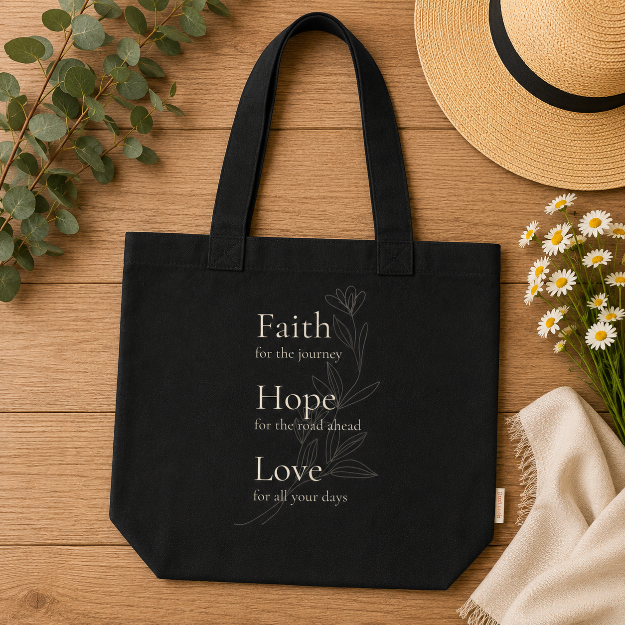 A black comfort colors canvas tote bag with white printed text reading 'Faith for the journey, Hope for the road ahead, Love for all your days' and a subtle botanical line drawing, styled on a wooden surface with eucalyptus leaves, a straw hat, daisies, and a cream cloth.