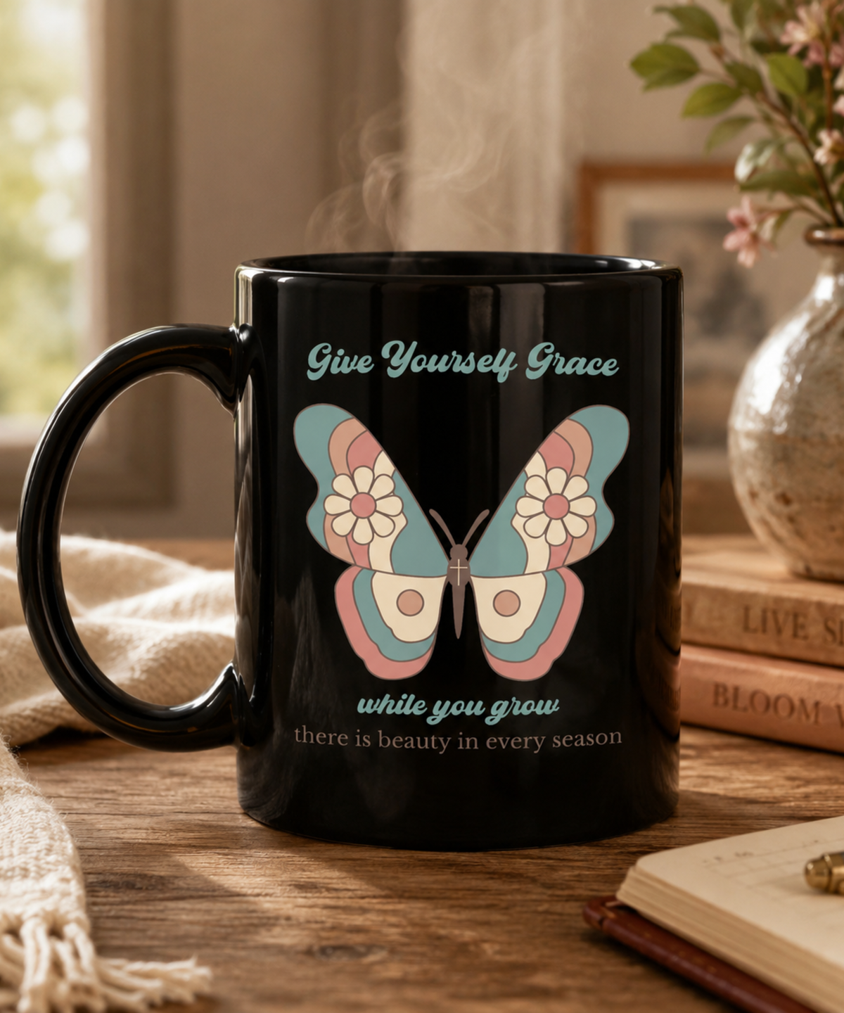 A black ceramic mug with a printed butterfly design and inspirational quote, give yourself grace while you grow. Styled on a wooden table with books and a vase in a warm indoor setting.