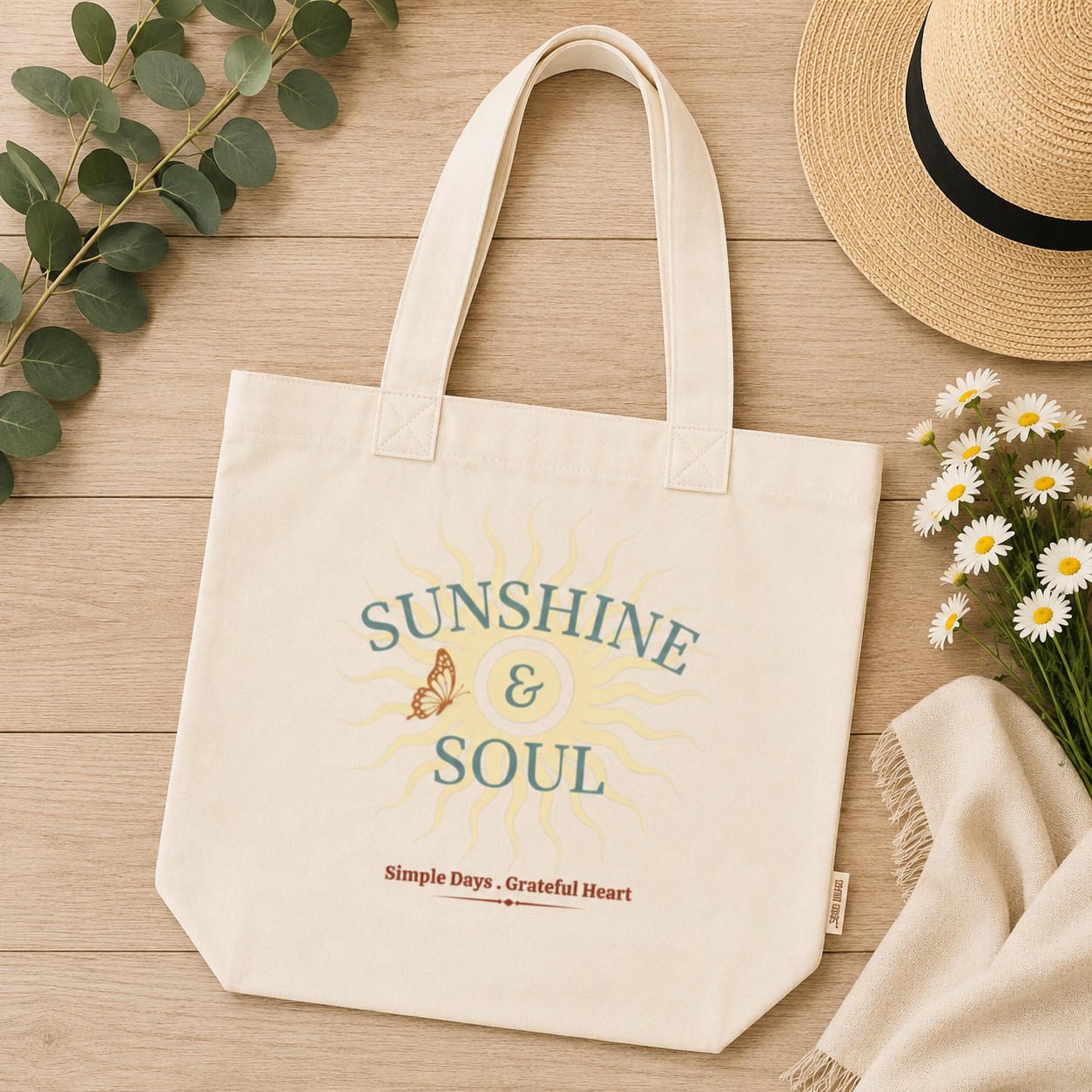 An Ivory comfort colors canvas tote bag with a 'SUNSHINE & SOUL' graphic print, styled in a flat-lay arrangement on a light wood background with a straw hat, daisies, eucalyptus, and a cream throw blanket.