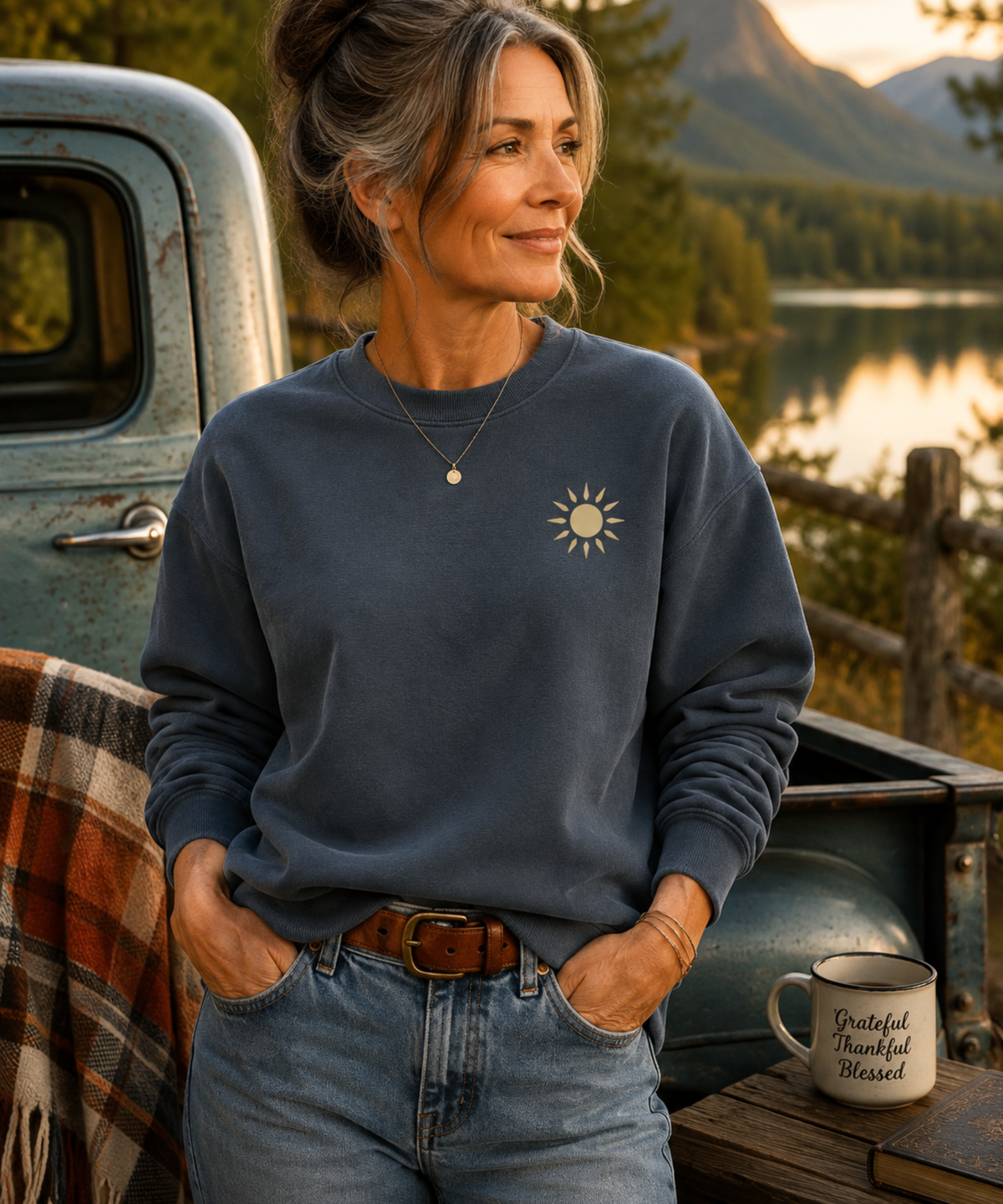 Woman wearing a denim blue garment-dyed crewneck sweatshirt with a small vintage sun graphic on the left chest outdoors by a mountain lake.