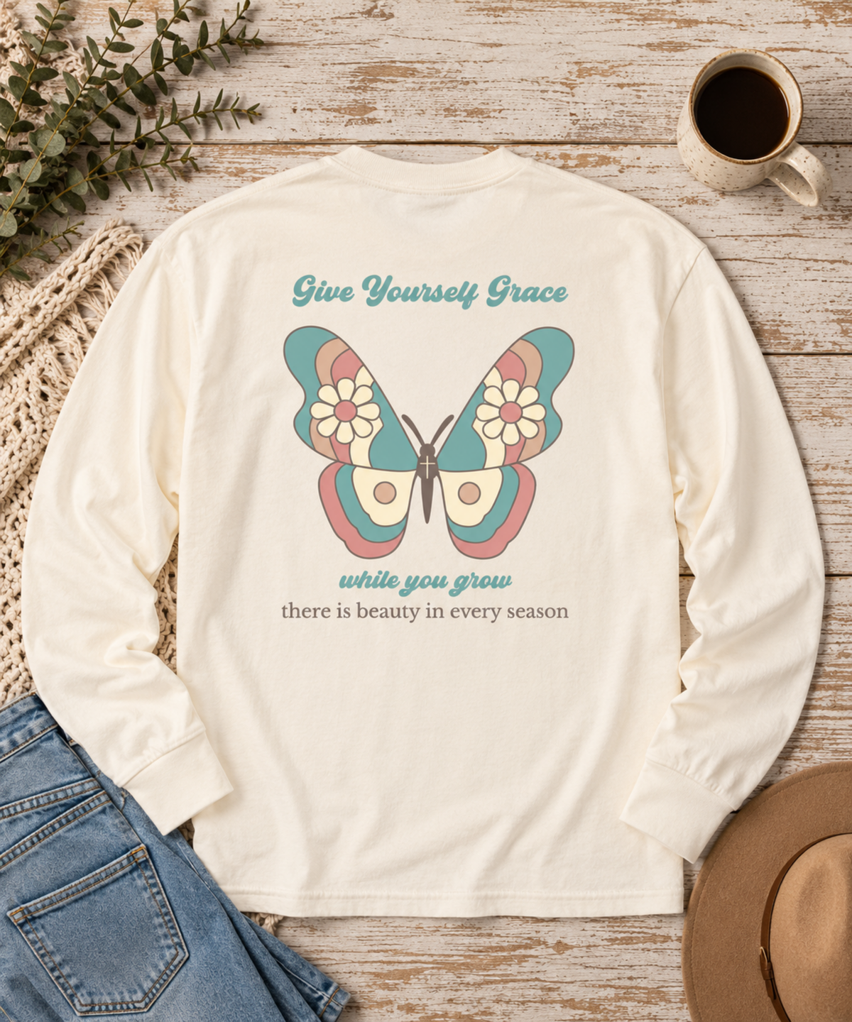 An Ivory long-sleeve shirt laid flat on a rustic wood surface, featuring a colorful vintage butterfly graphic and the text 'Give Yourself Grace while you grow there is beauty in every season.' Jeans, a coffee cup, a hat, and greenery are styled around it.