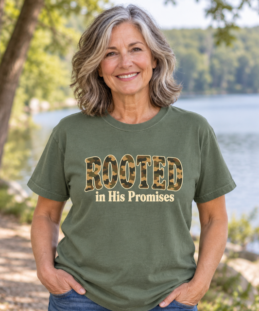 A mature women wearing a sage green Comfort Colors tee with Rooted in His Promises camo lettering and cream text.