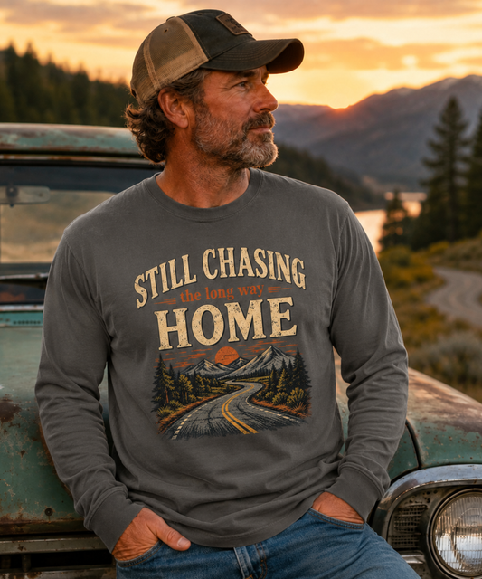 A man wearing a pepper long-sleeve graphic t-shirt with the words Still Chasing the long way Home and a trucker cap, leaning against a vintage green car with a mountain lake sunset background.