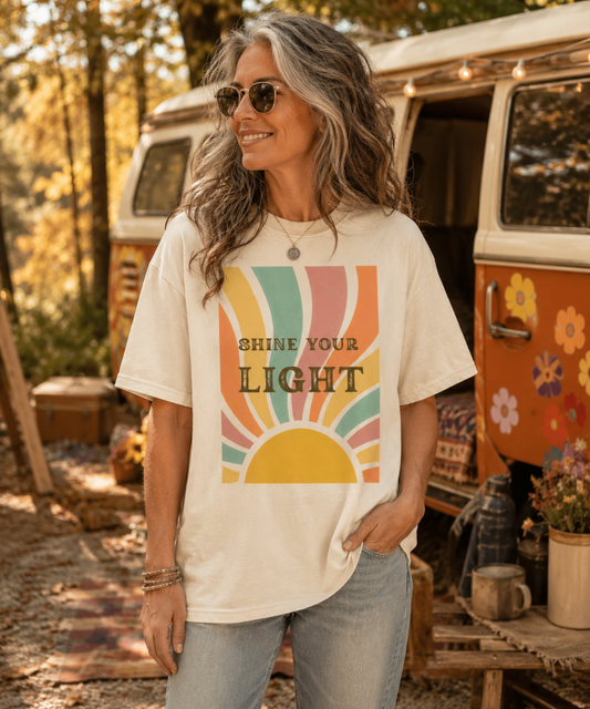 A woman wearing an ivory comfort-colors graphic t-shirt with a retro sunset design and the text 'SHINE YOUR LIGHT', standing in front of a vintage camper van in an outdoor campsite setting.