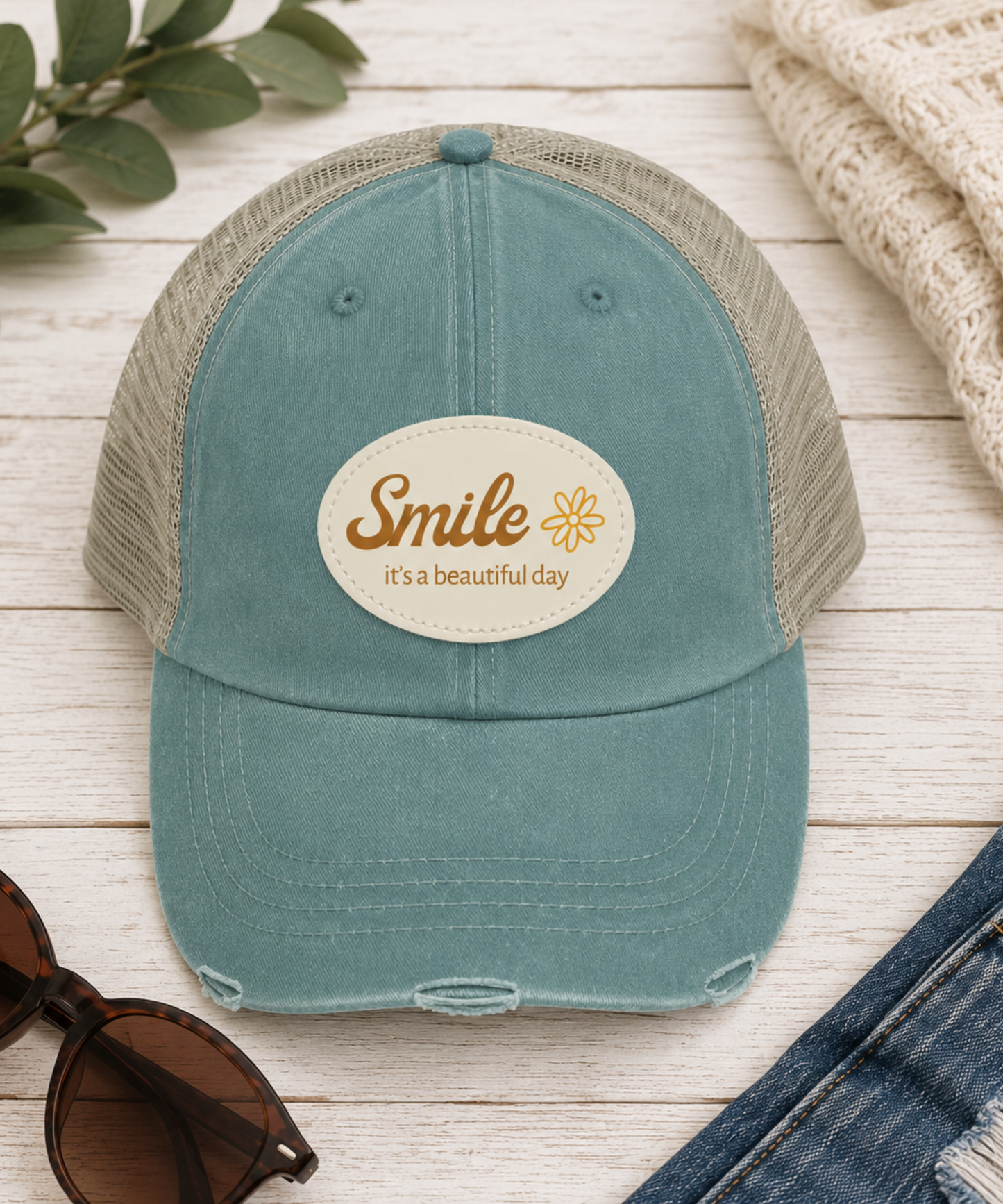 A forest trucker hat with a tan mesh back and a cream oval patch reading 'Smile it's a beautiful day' with a small flower graphic. The hat is styled in a flat-lay on a white wood background with sunglasses, a knit sweater, denim shorts, and green leaves.