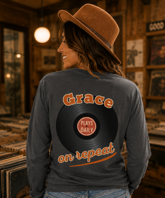 A woman wearing a brown wide-brim hat and a pepper comfort colors long-sleeve shirt with a graphic on the back. The graphic features the word 'Grace' above a vinyl record with 'PLAYS DAILY' in the center, and 'on repeat' below in orange.