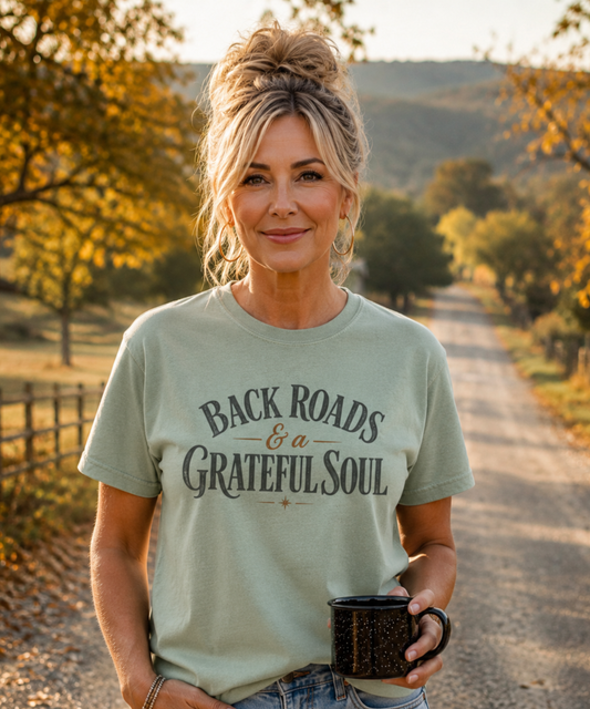 A woman stands on a rural dirt road wearing a bay green distress graphic t-shirt that reads 'BACKROADS & A GRATEFUL SOUL' in dark gray lettering. She is holding a black speckled mug, with trees and hills in the background.