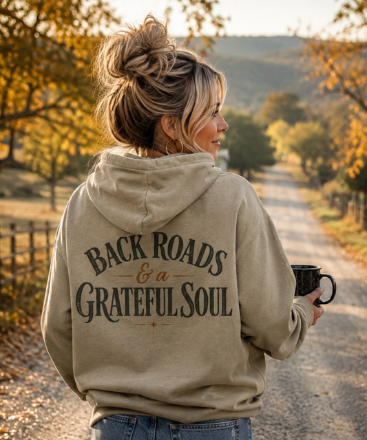 A woman stands on a country road wearing a gray comfort colors hoodie from the back, which features the distressed text 'BACKROADS & A GRATEFUL SOUL' in dark lettering. She is holding a black mug, with trees and hills in the background.