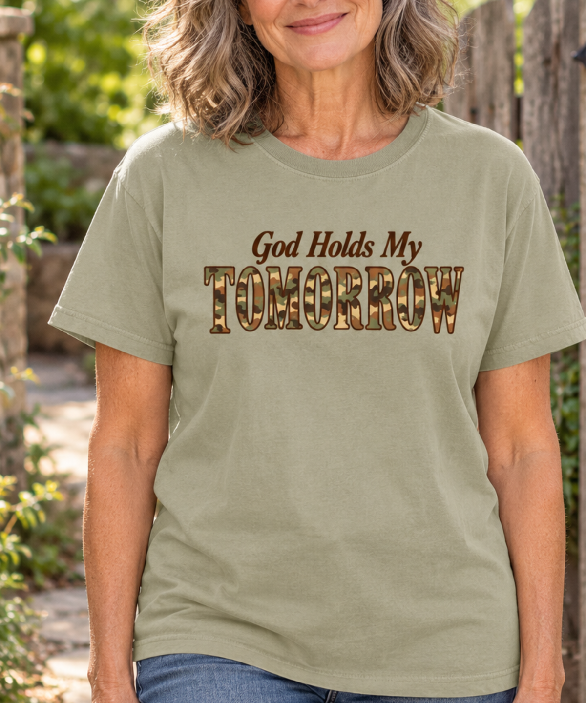 Woman with salt-and-pepper hair wearing a khaki God Holds My Tomorrow camo lettering tee and jeans beside a wooden garden gate.