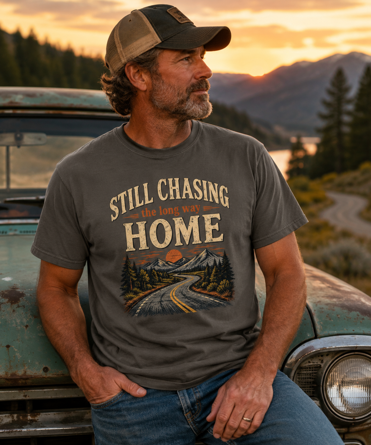 A man wearing a pepper t-shirt with a mountain road print and the text 'STILL CHASING the long way HOME', standing outdoors next to a vintage car with mountains in the background.