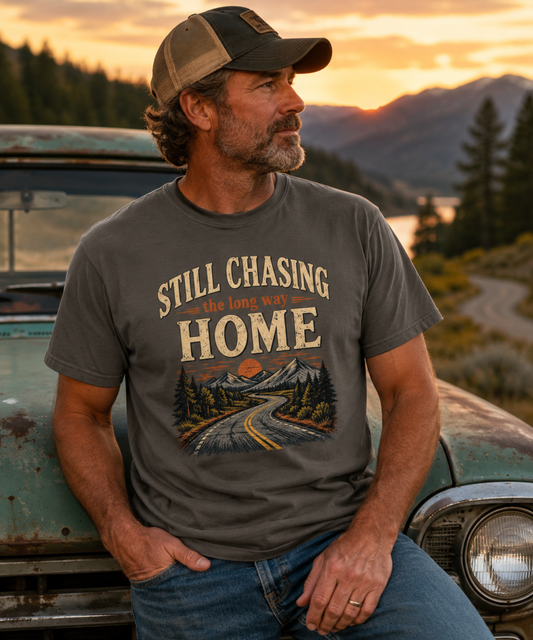 A man wearing a pepper t-shirt with a mountain road print and the text 'STILL CHASING the long way HOME', standing outdoors next to a vintage car with mountains in the background.