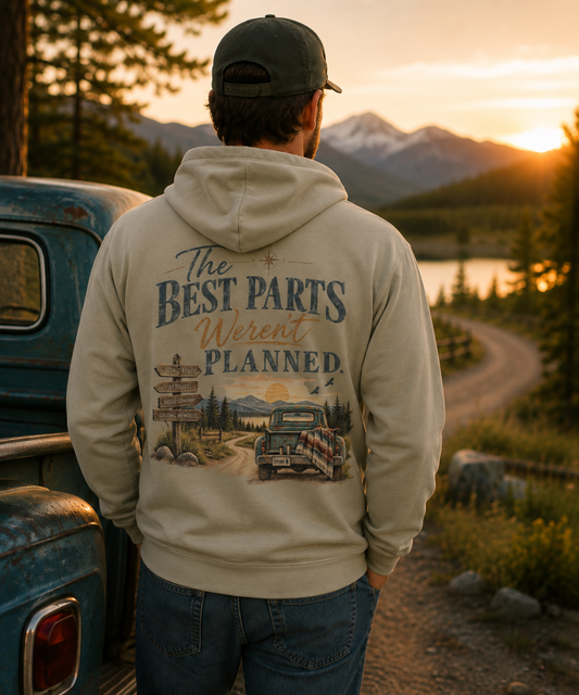 A man stands with his back to the camera wearing a gray hoodie with a large vintage-style back graphic that reads 'The Best Parts Weren't Planned.' He is outdoors at sunset near a lake, mountains, trees, and a blue pickup truck.