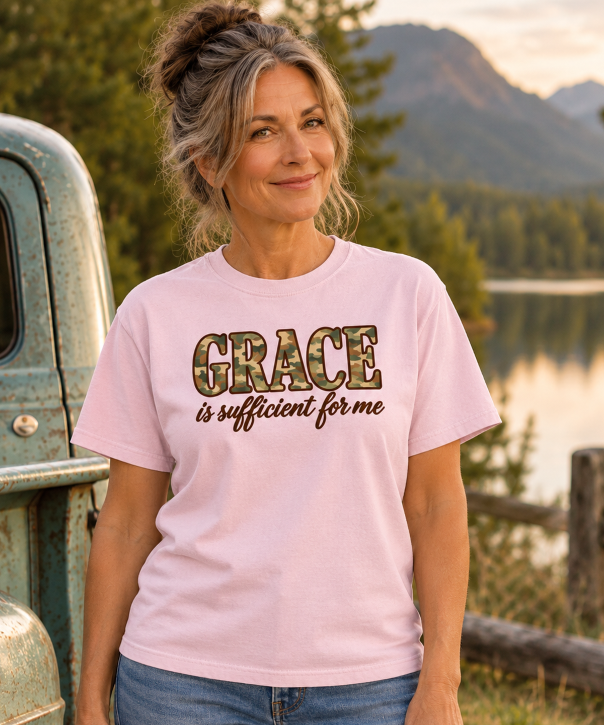 A woman wearing a light pink graphic t-shirt with the text 'GRACE is sufficient for me' camo and brown, standing outdoors next to a vintage truck with a mountain and lake background.