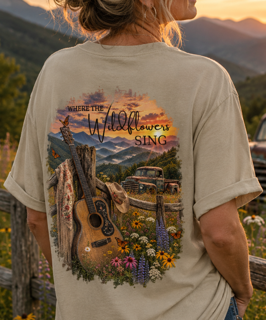 A woman shown from behind wearing a sandstone comfort colors graphic t-shirt with a mountain scene print. The print features an acoustic guitar, a rustic truck, wildflowers, and the text 'WHERE THE Wildflowers SING'.