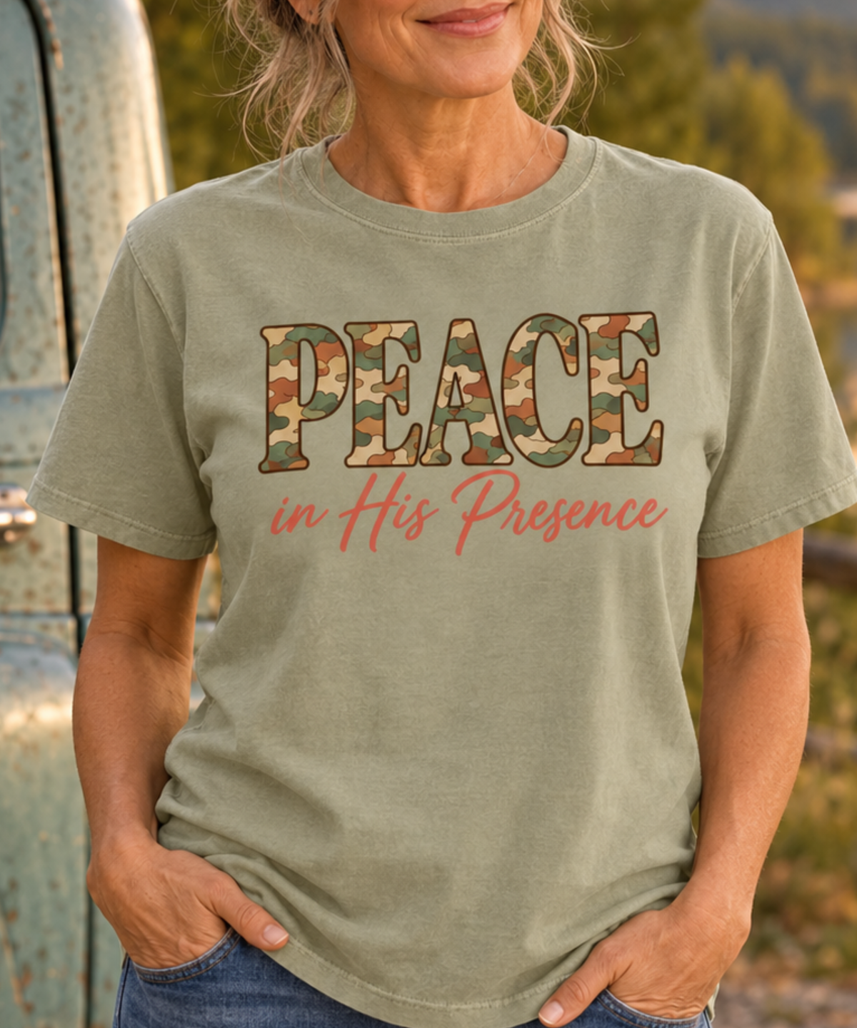 A woman wearing a khaki graphic t-shirt with 'PEACE in His Presence' printed on the front. The word 'PEACE' is filled with a camouflage pattern, and 'in His Presence' is written in a coral pink script.