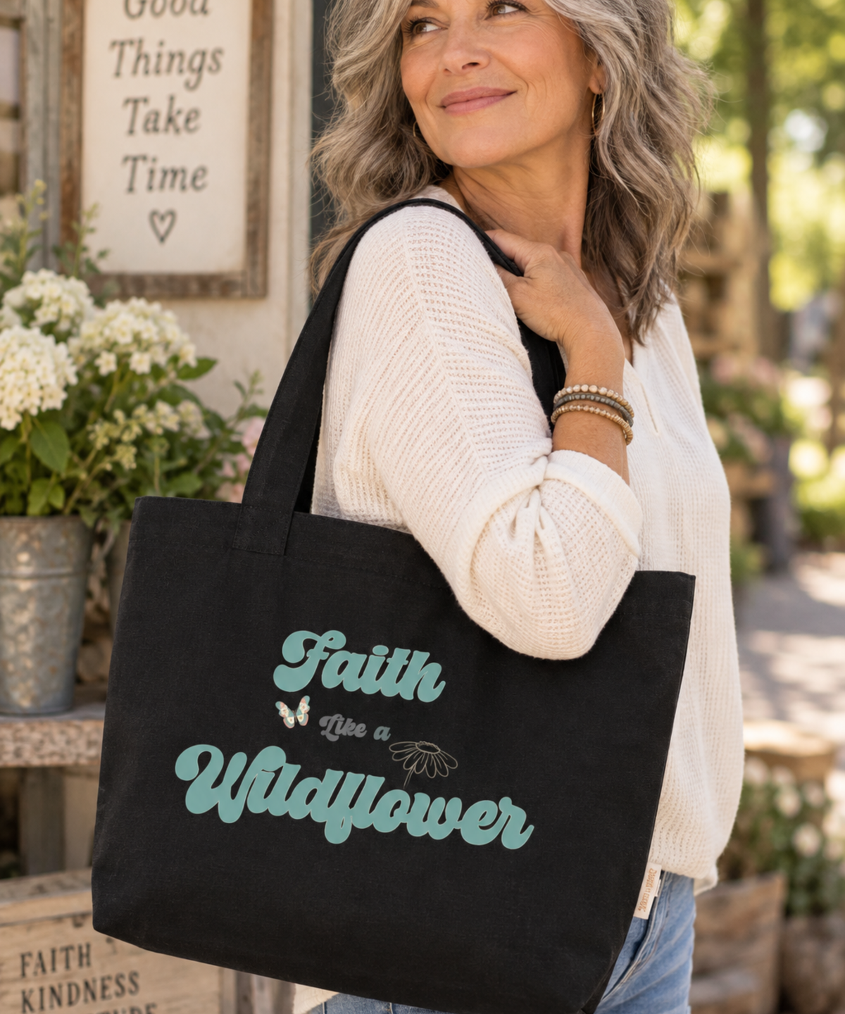 A woman wearing a cream knit sweater and blue jeans carries a black canvas tote bag with the phrase 'Faith Like a Wildflower' in teel script and small butterfly/leaf illustrations. The photo is taken in a softly blurred outdoor shop or garden setting.
