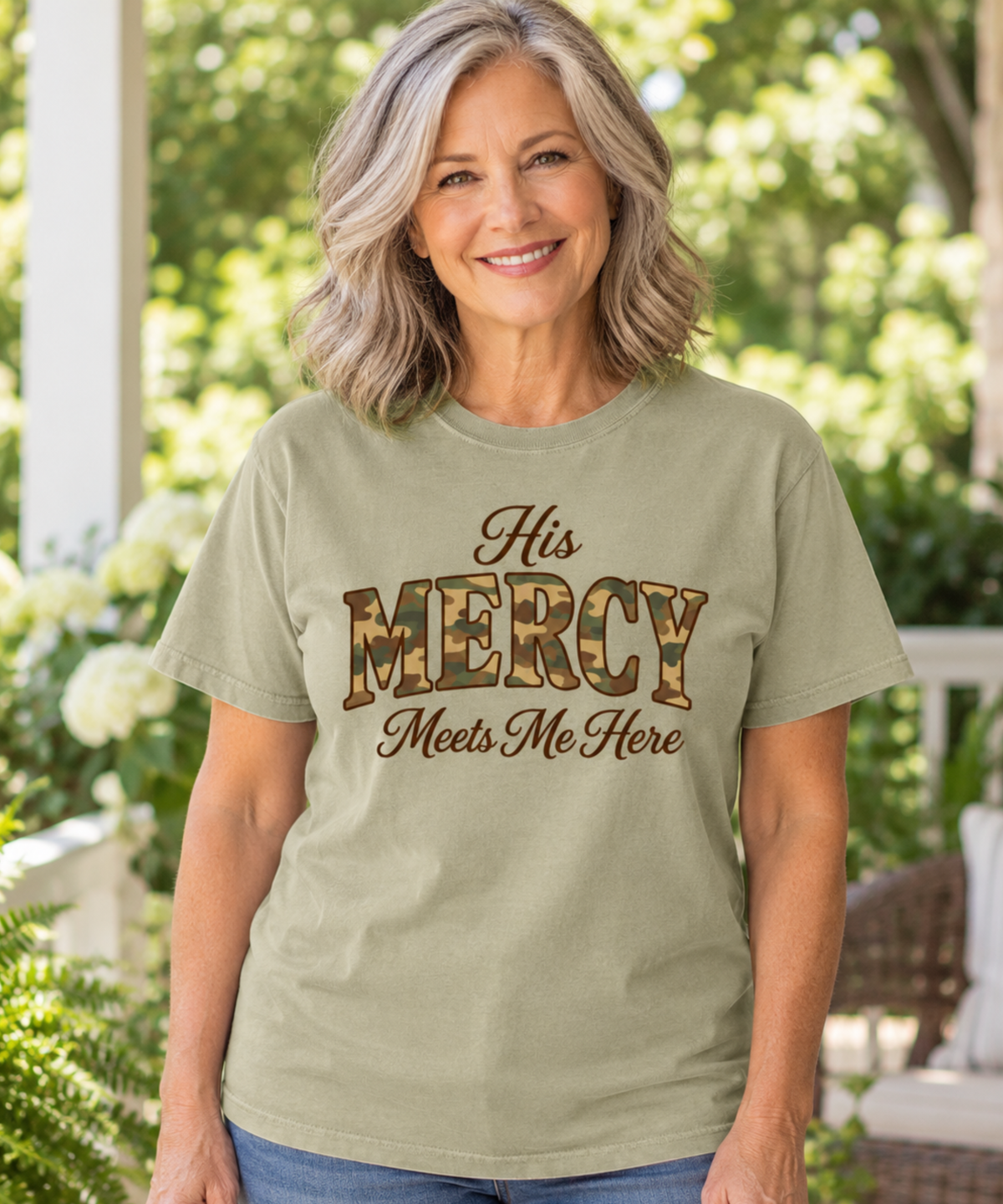 A smiling woman with shoulder-length gray hair stands outdoors wearing a khaki graphic T-shirt with the words 'His MERCY Meets Me Here' printed on the front. The word 'MERCY' is styled in a camo print pattern.