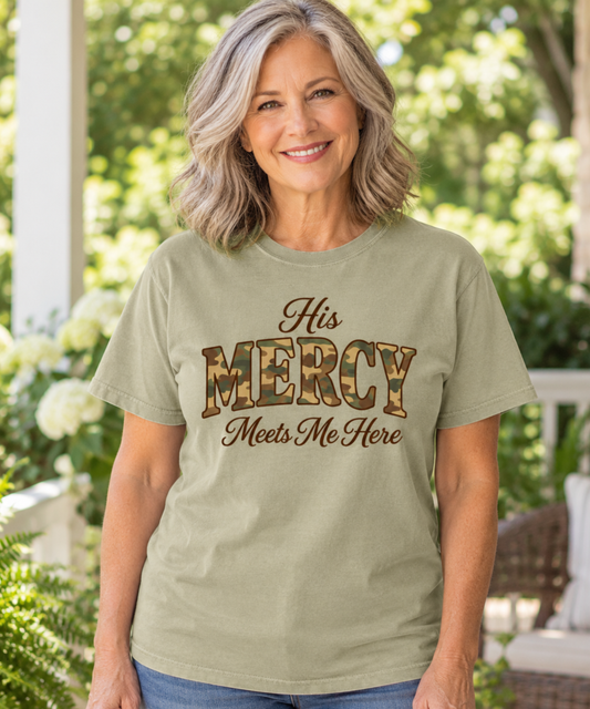A smiling woman with shoulder-length gray hair stands outdoors wearing a khaki graphic T-shirt with the words 'His MERCY Meets Me Here' printed on the front. The word 'MERCY' is styled in a camo print pattern.