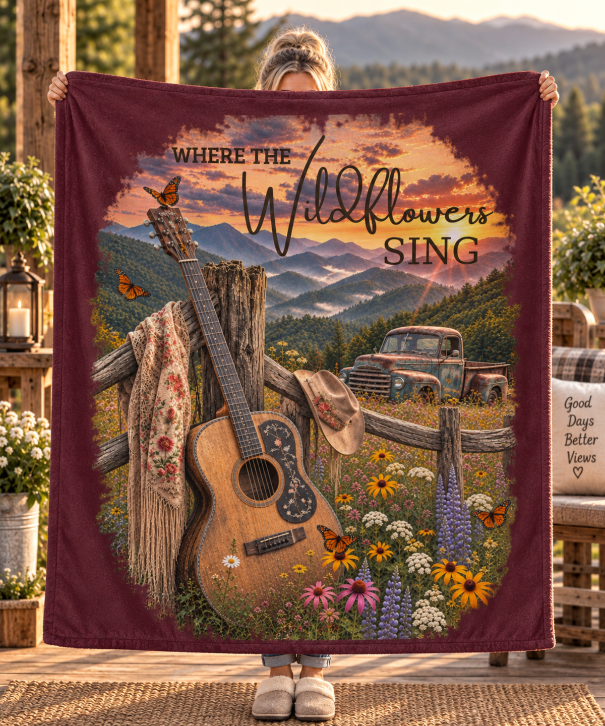 A person holds up a large rectangular blanket or throw featuring a rustic mountain landscape scene with the text 'WHERE THE Wildflowers SING.' The blanket has a maroon border and a printed design including an acoustic guitar, fence, flowers, butterflies, a truck, and mountains.