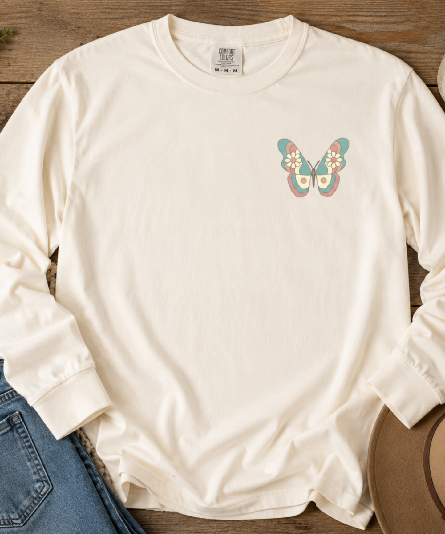 An Ivory comfort colors long-sleeve crewneck t-shirt with a colorful vintage butterfly graphic on the left chest, styled flat on a wooden surface next to blue jeans and a brown hat.