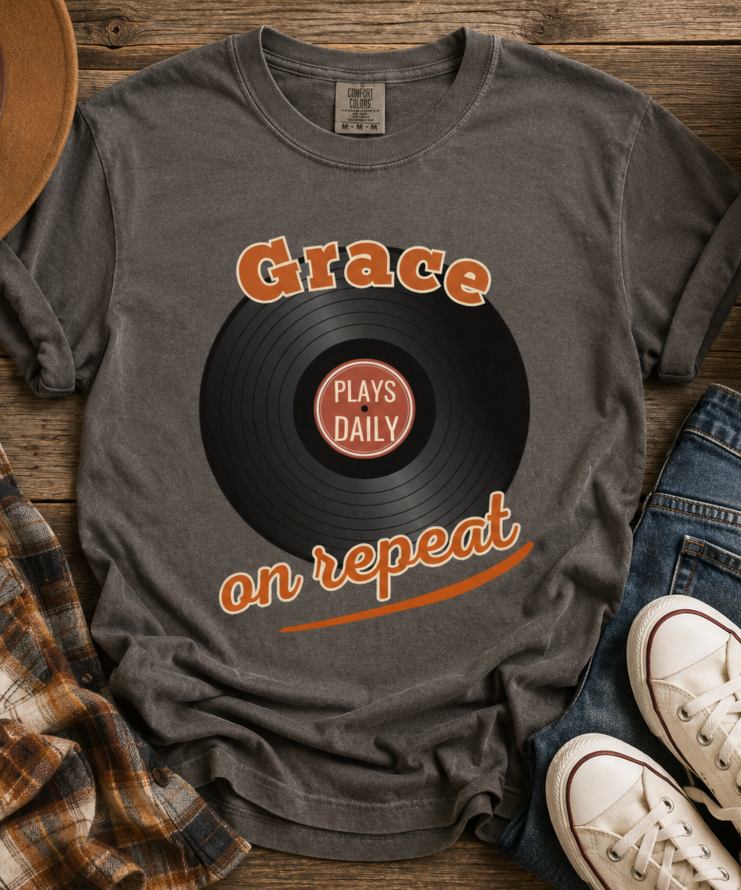A pepper comfort colors graphic t-shirt laid flat on a rustic wooden surface, styled with a plaid shirt, jeans, white sneakers, and a hat. The t-shirt features a black vinyl record graphic with the words 'Grace on repeat' in orange and 'PLAYS DAILY' in white in the center.