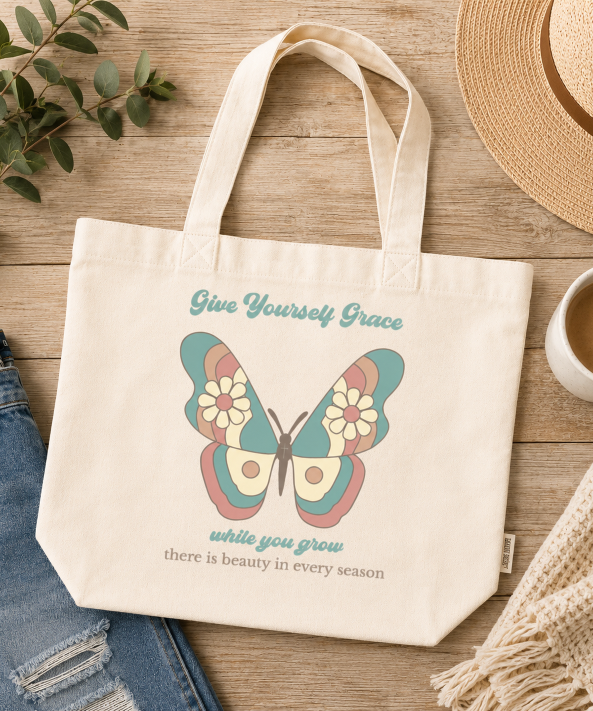 An Ivory comfort colors canvas tote bag with a colorful butterfly graphic and the text 'Give Yourself Grace while you grow there is beauty in every season', styled on a wooden surface with jeans, a straw hat, a cup of coffee, and a knit scarf.