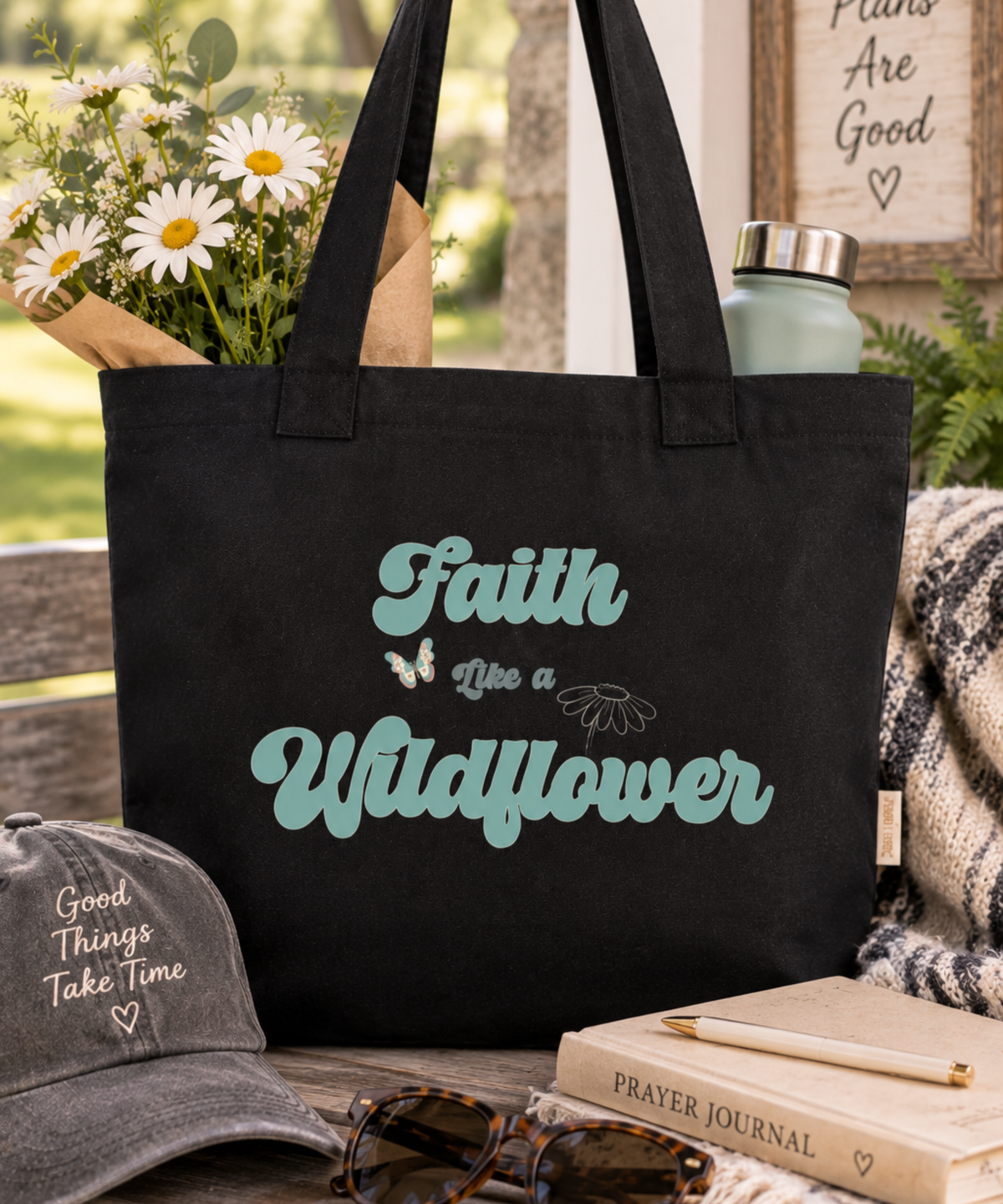 A black canvas tote bag with the phrase 'Faith Like a Wildflower' in teal lettering, styled in a rustic outdoor setting with flowers, a gray baseball cap, sunglasses, a prayer journal, and a notebook.