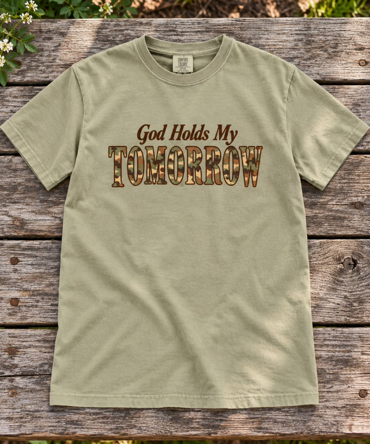 Khaki Comfort Colors tee with God Holds My Tomorrow in brown and camo lettering, laid flat on an outdoor wooden table surrounded by greenery.