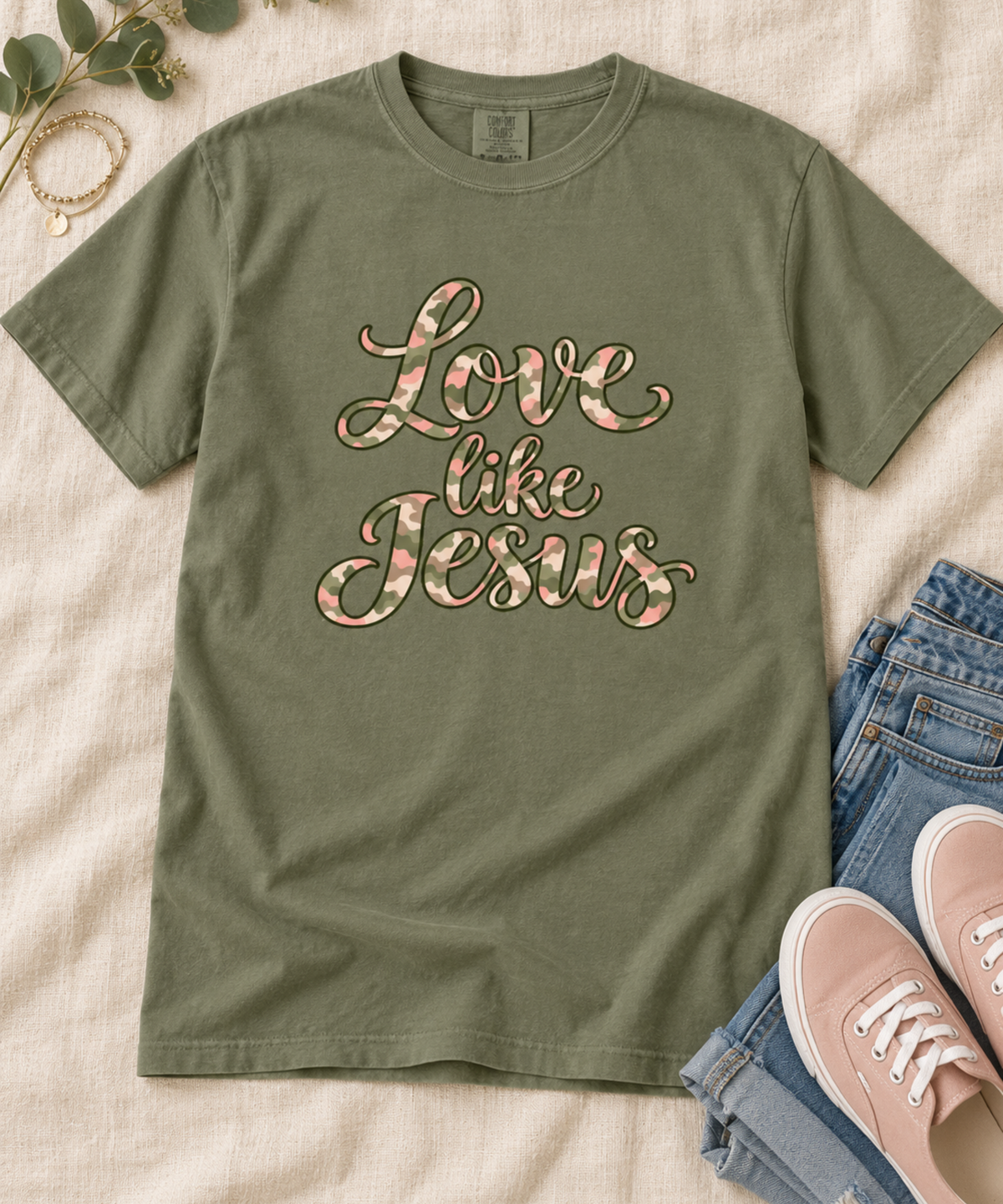 A moss green short-sleeve t-shirt with 'Love like Jesus' printed in a pink, cream, and green camo with script across the chest, styled in a flat lay with blue jeans, pink sneakers, and a gold necklace on a beige fabric background.