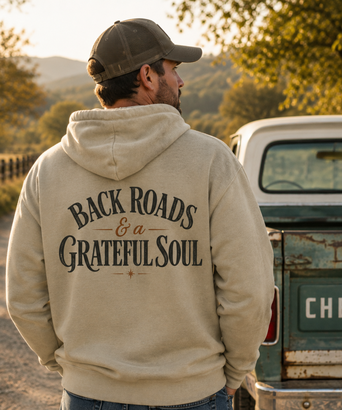 A man wearing a gray comfort colors hoodie with distressed 'BACKROADS & a GRATEFUL SOUL' printed on the back, standing next to a vintage truck in an outdoor setting during golden hour.