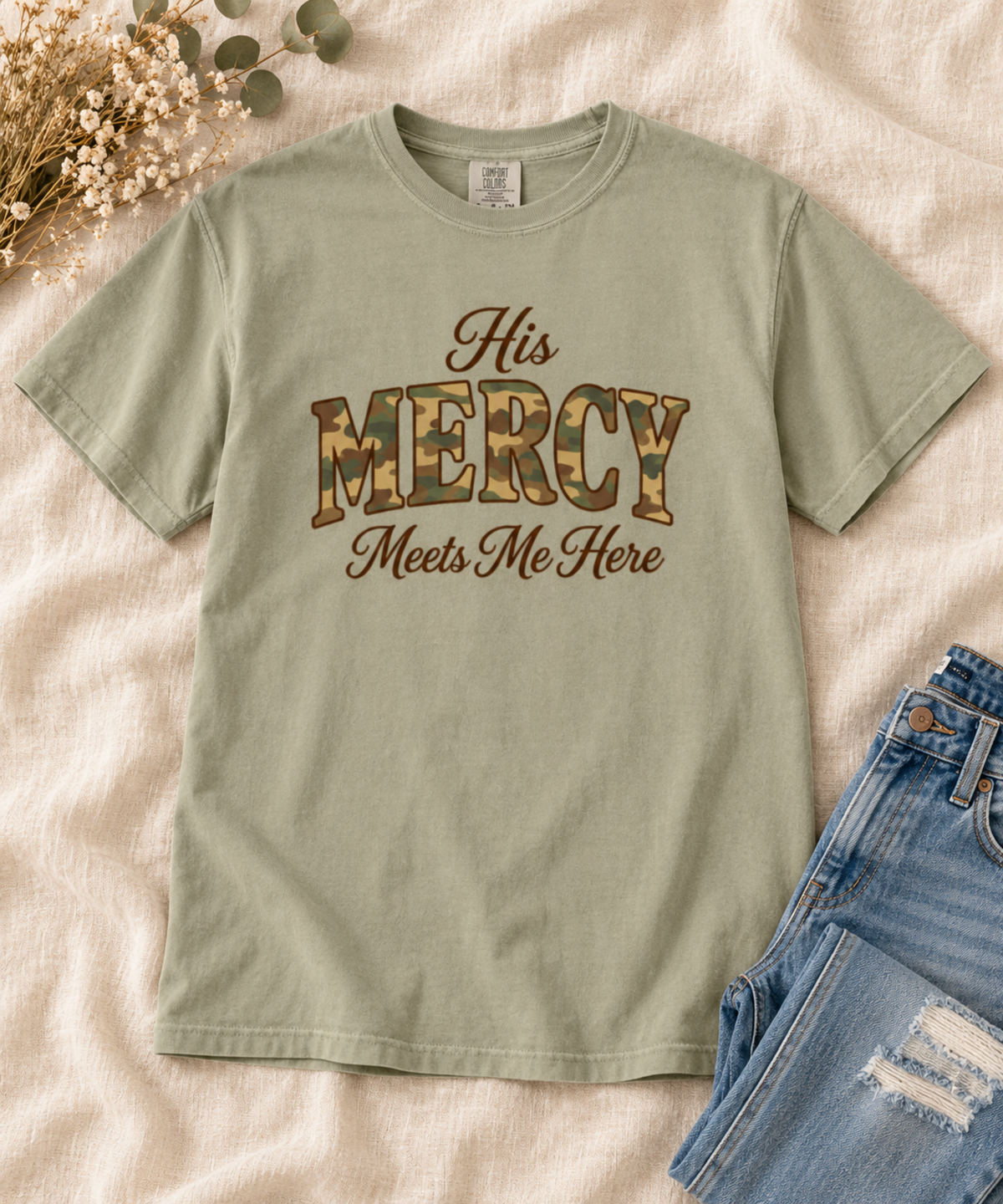A khaki short-sleeve comfort colors t-shirt with a graphic reading 'His MERCY Meets Me Here' in brown and camouflage lettering, styled flat-lay on a beige fabric background with dried flowers and blue jeans.