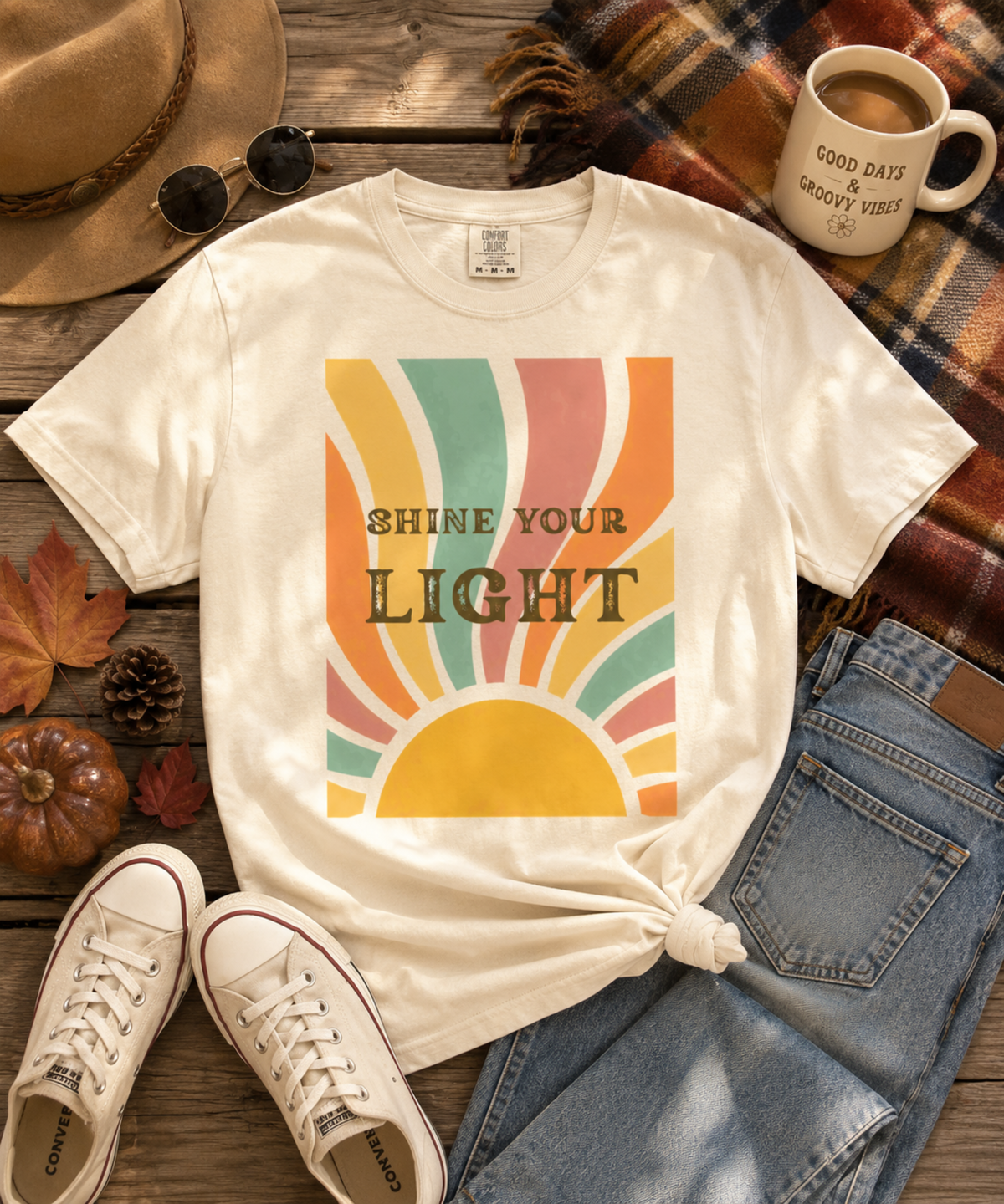 An ivory comfort colors graphic t-shirt styled in a flat lay with a tie knot, surrounded by a tan hat, sunglasses, a coffee mug, blue jeans, white sneakers, and autumn leaves on a rustic wooden background. The t-shirt features a retro sunburst graphic with the text 'SHINE YOUR LIGHT'.