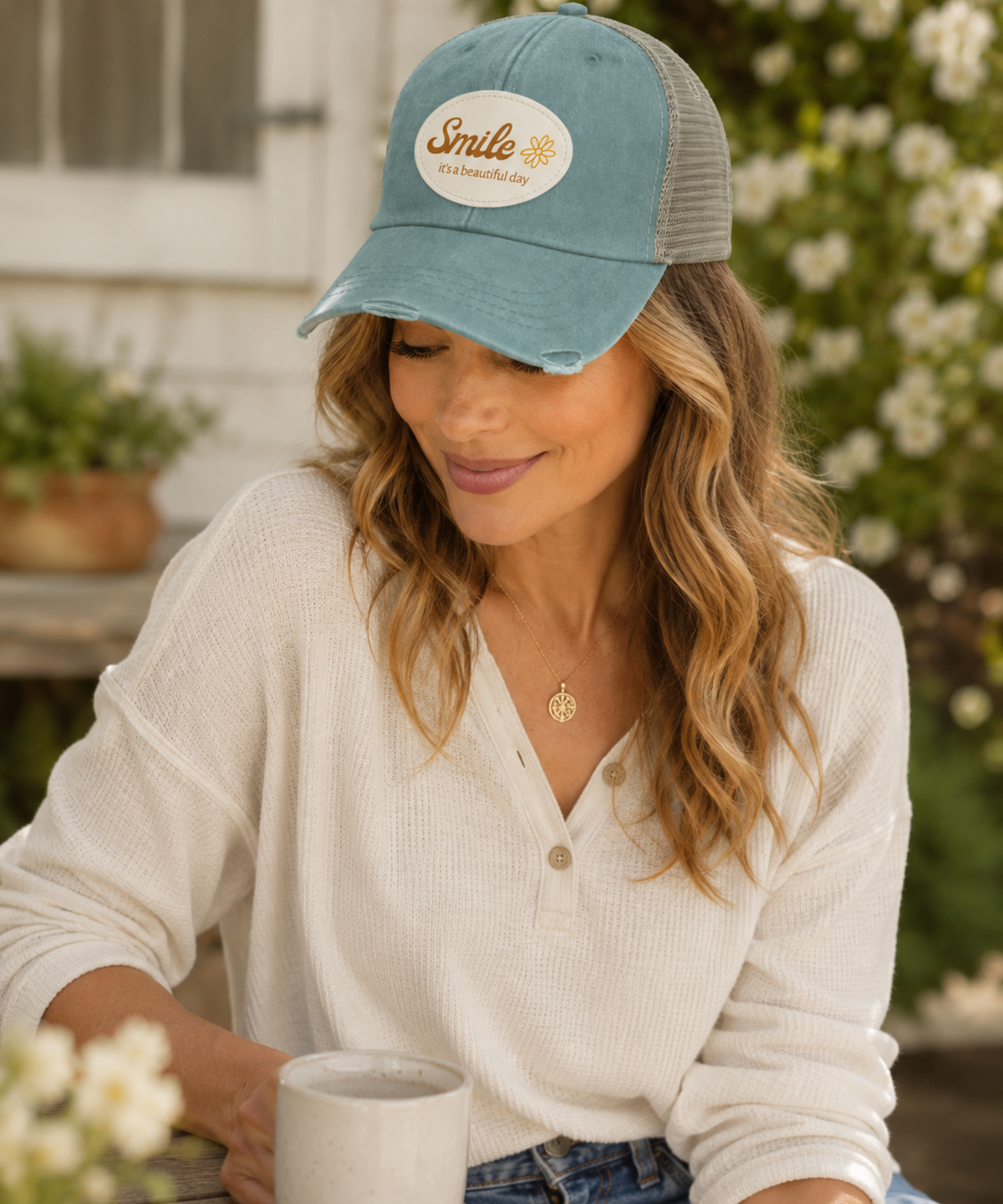 A woman wearing a distressed forest trucker hat with a mesh back and a oval 'Smile with Sunshine' patch on the front, sitting outdoors and holding a mug.