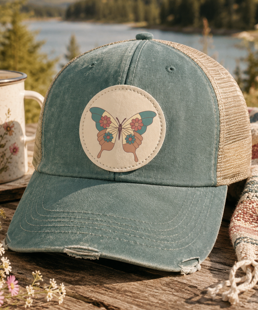Forest Vintage butterfly patch trucker hat displayed on a rustic wooden table with a camping mug, woven blanket, and wildflowers overlooking a peaceful lake and evergreen forest.