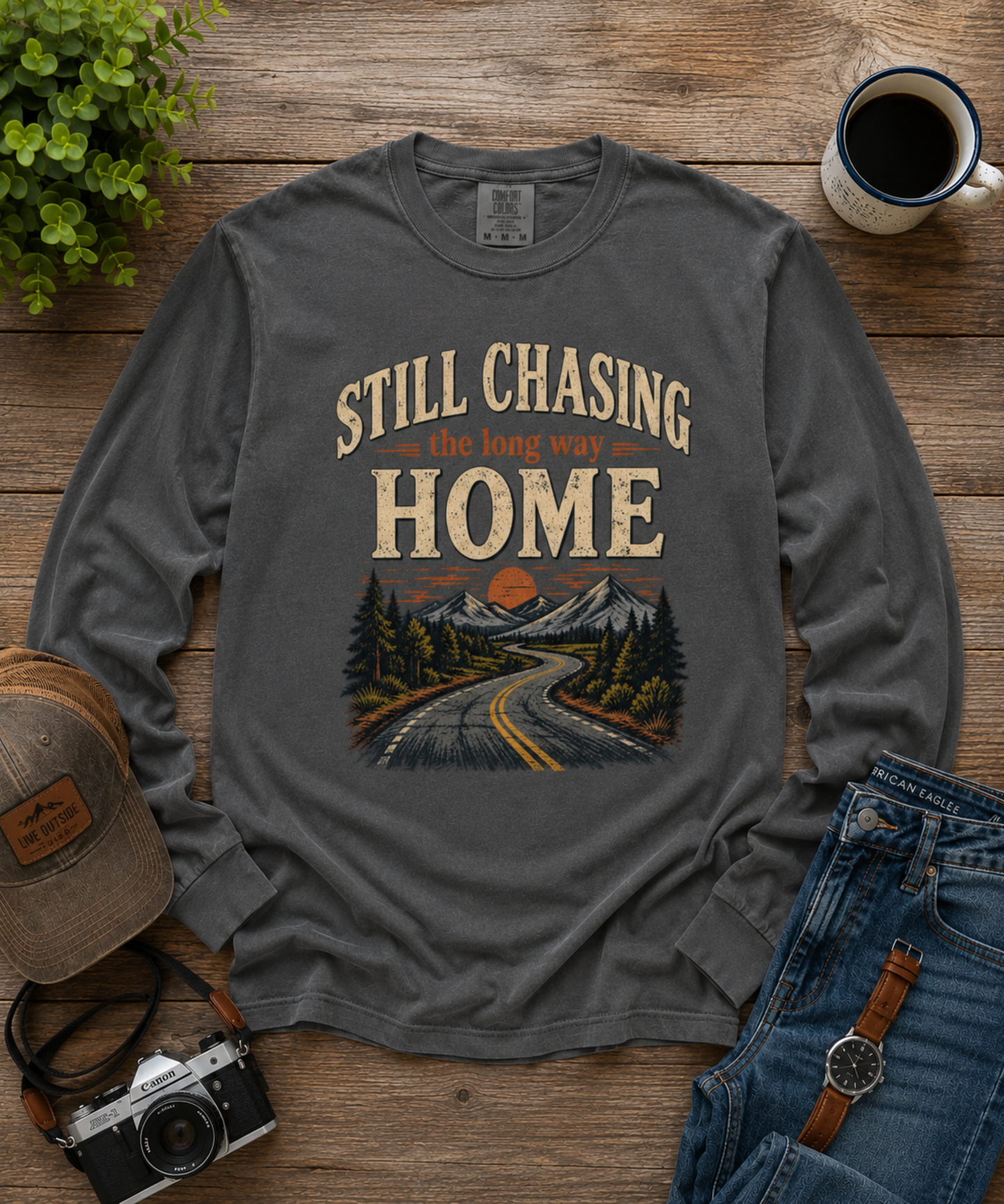 A pepper colored long-sleeve graphic t-shirt laid flat on a rustic wooden surface, surrounded by lifestyle props including a vintage-style camera, a cap, blue jeans, a watch, a mug of coffee, and a small green plant. The shirt features a graphic print of a winding mountain road with the text 'STILL CHASING the long way HOME'.