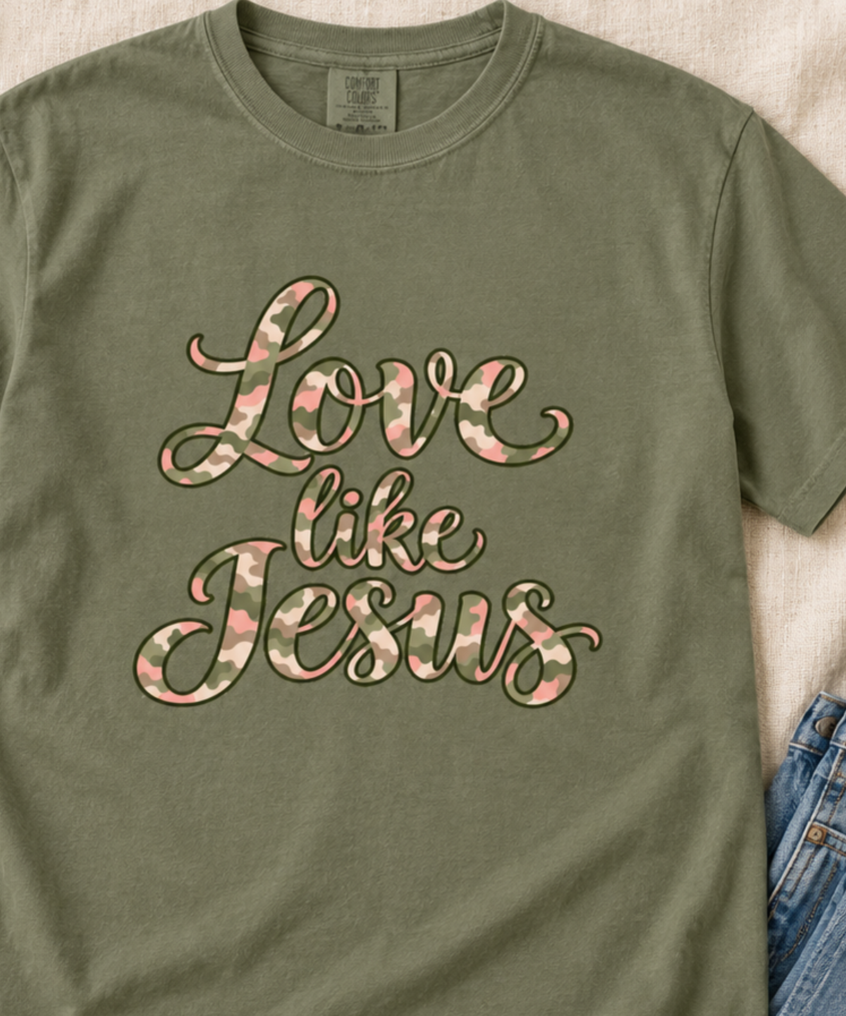Close up of a moss green comfort colors t-shirt featuring the phrase 'Love like Jesus' in a cursive script filled with a pink, cream and green camouflage pattern, laid flat on a light beige fabric background with a pair of blue jeans partially visible on the right.