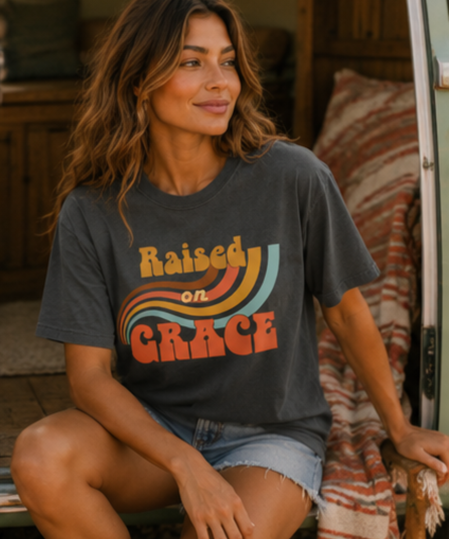 A woman wearing a graphite comfort colors graphic t-shirt with a retro rainbow design and the text 'Raised on Grace'. She is sitting in a cozy, rustic indoor setting with denim shorts.