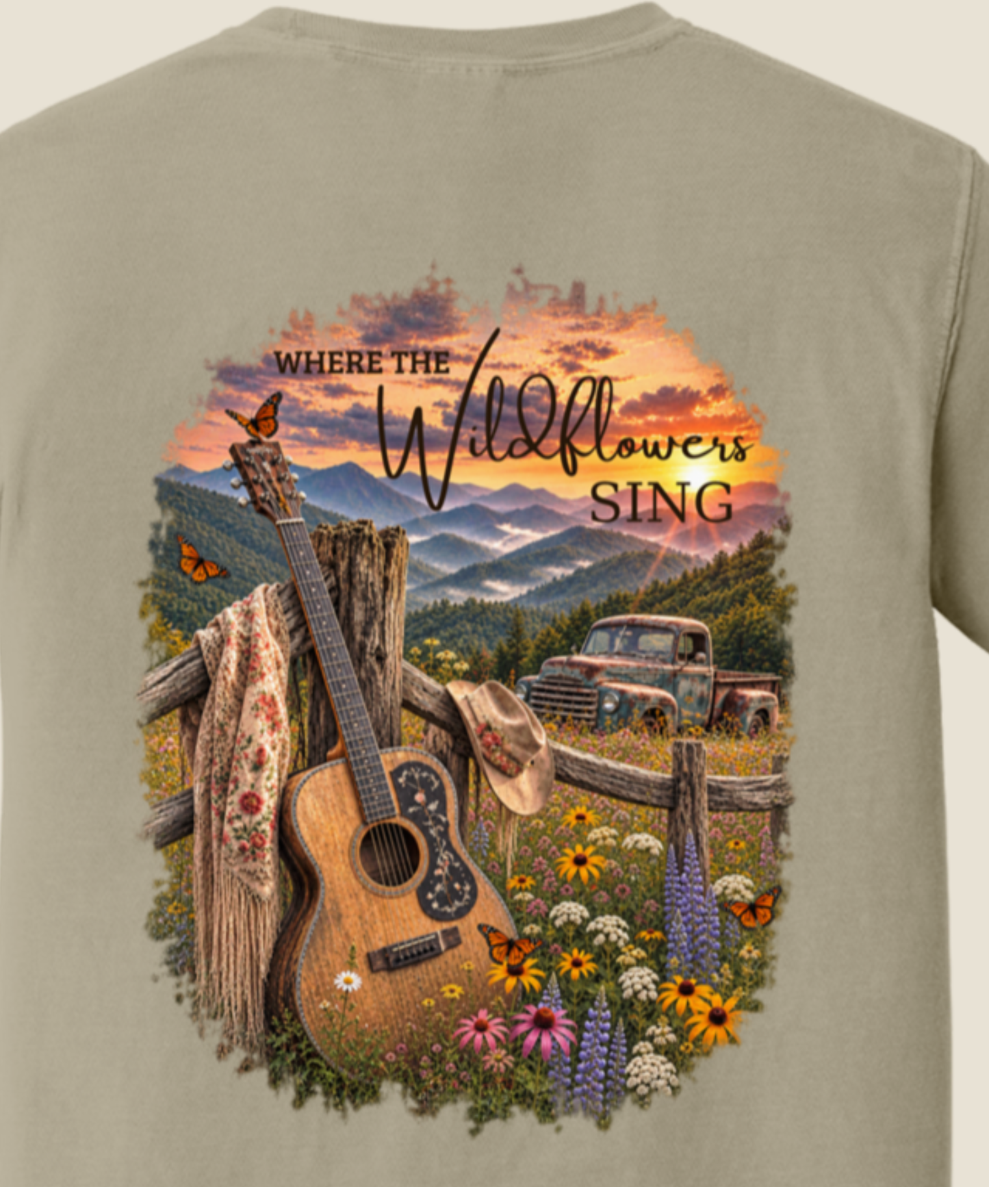 A close-up of the back of a sandstone t-shirt featuring a large, colorful graphic print. The graphic depicts a rustic country scene with a wooden fence, a guitar leaning on a post, a cowboy hat, wildflowers, a vintage pickup truck, and mountains under a sunset sky, with the text 'WHERE THE Wildflowers SING'.