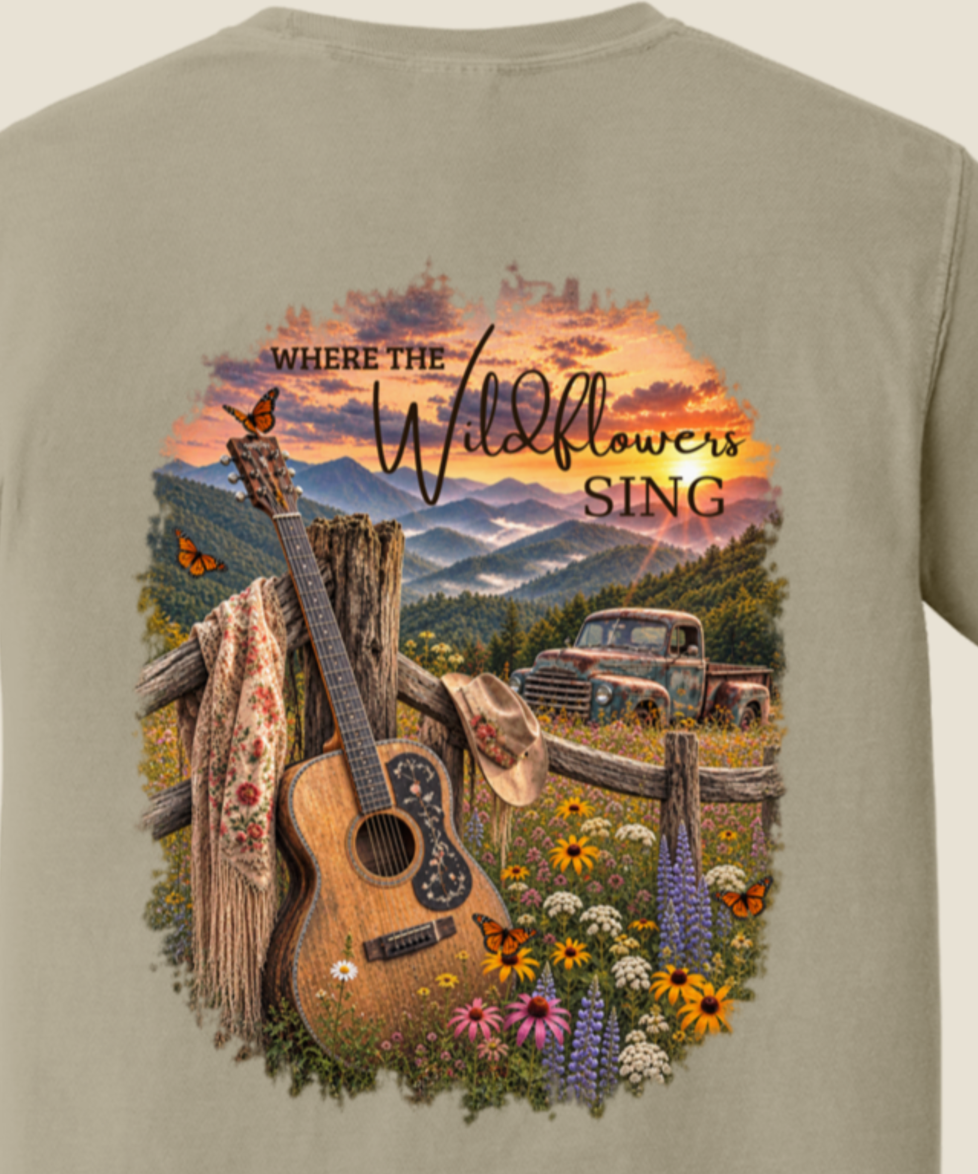 A close-up of the back of a sandstone t-shirt featuring a large, colorful graphic print. The graphic depicts a rustic country scene with a wooden fence, a guitar leaning on a post, a cowboy hat, wildflowers, a vintage pickup truck, and mountains under a sunset sky, with the text 'WHERE THE Wildflowers SING'.