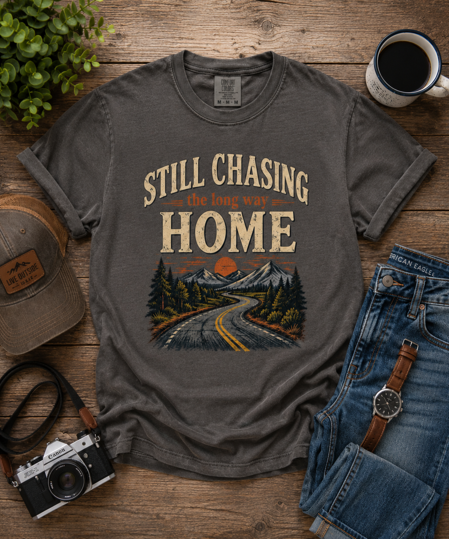 A pepper graphic t-shirt laid flat on a rustic wooden surface, surrounded by lifestyle props including a vintage camera, a baseball cap, folded blue jeans, a watch, a mug of coffee, and a small plant. The t-shirt features a retro-style graphic with the text 'STILL CHASING the long way HOME' above an illustration of a winding road through mountains and pine trees.