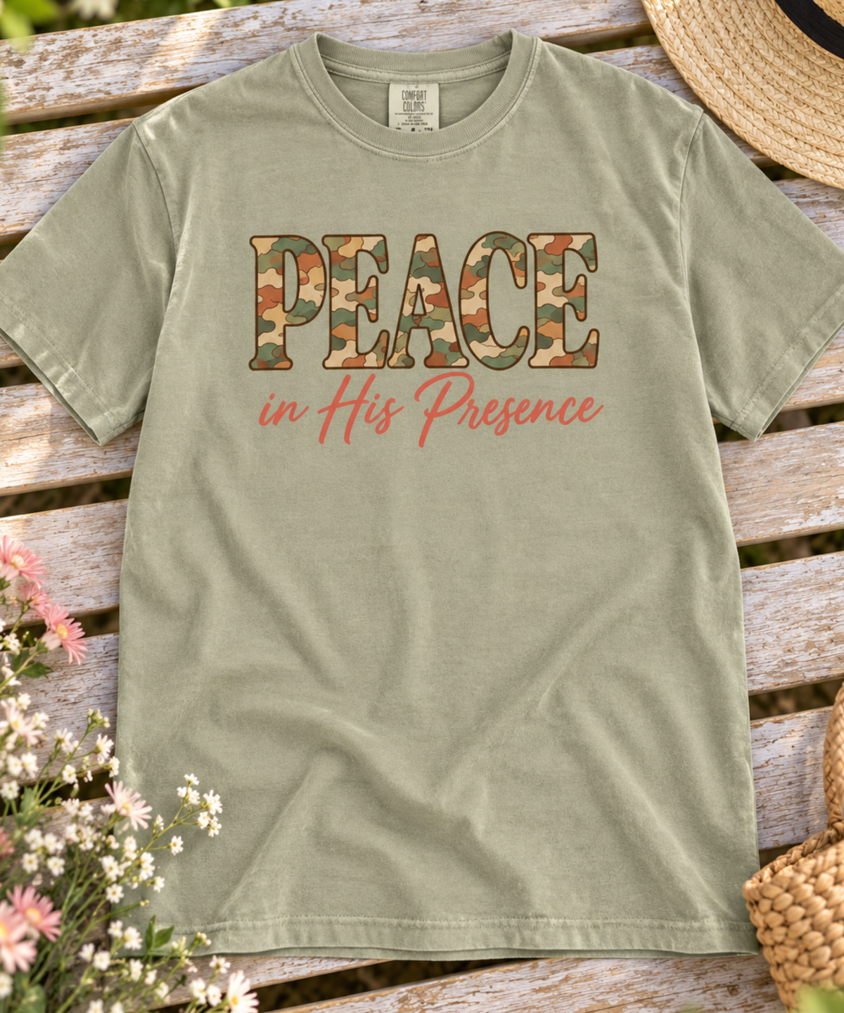 A khaki comfort colors t-shirt laid flat on a rustic wooden bench, featuring the text 'PEACE in His Presence' in camouflage and coral script. Decorative flowers and a straw hat frame the edges of the shot.
