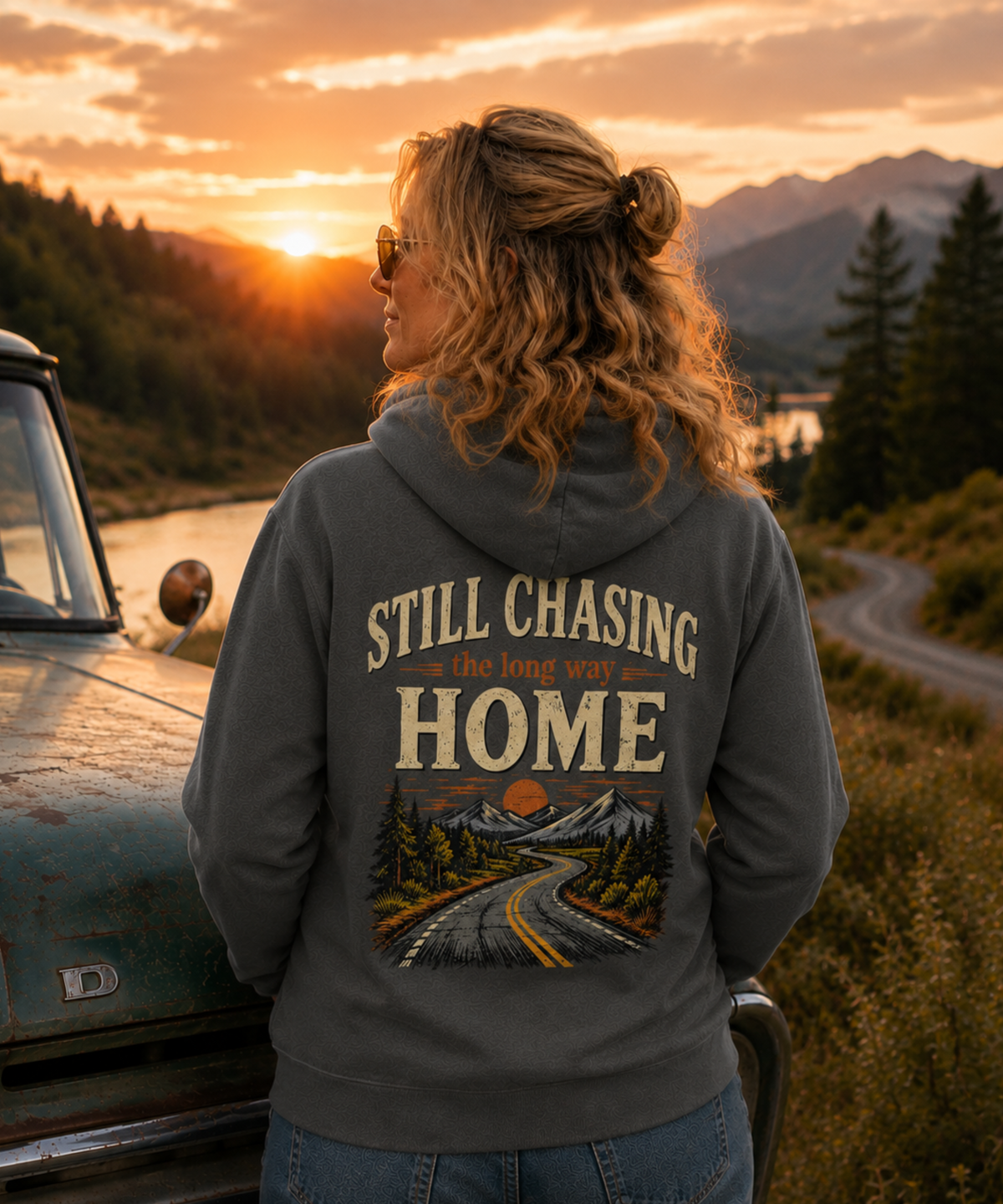 A woman stands outdoors at sunset beside a vintage truck, wearing a pepper colored hoodie with a large back graphic that reads “Still Chasing the Long Way Home” above a mountain road illustration.