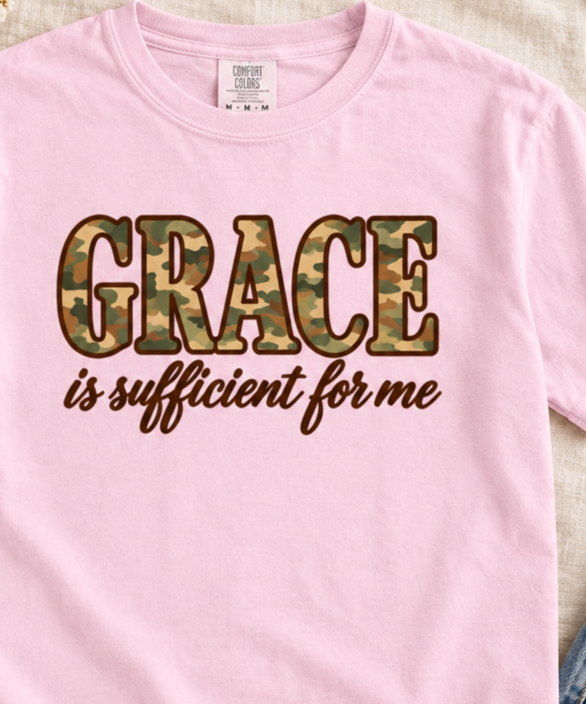 Close up of a light pink Comfort Colors t-shirt featuring the text 'GRACE is sufficient for me' printed on the front. The word 'GRACE' is styled in a green and brown camouflage pattern, while 'is sufficient for me' is written below in a dark brown cursive font.