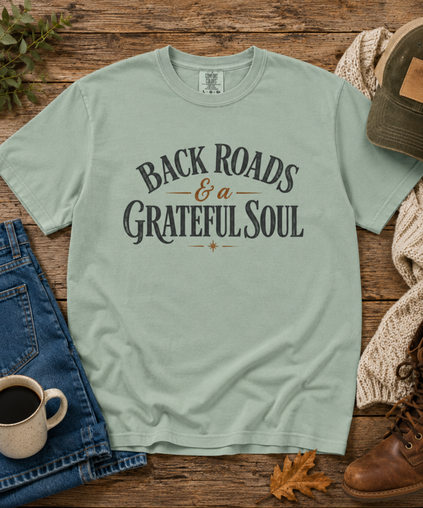 A bay green distressed graphic t-shirt laid flat on a rustic wooden surface, surrounded by lifestyle props including blue jeans, a coffee mug, a knit sweater, a brown leather boot, and a baseball cap. The t-shirt features the text 'BACK ROADS & a GRATEFUL SOUL' in dark gray and brown.