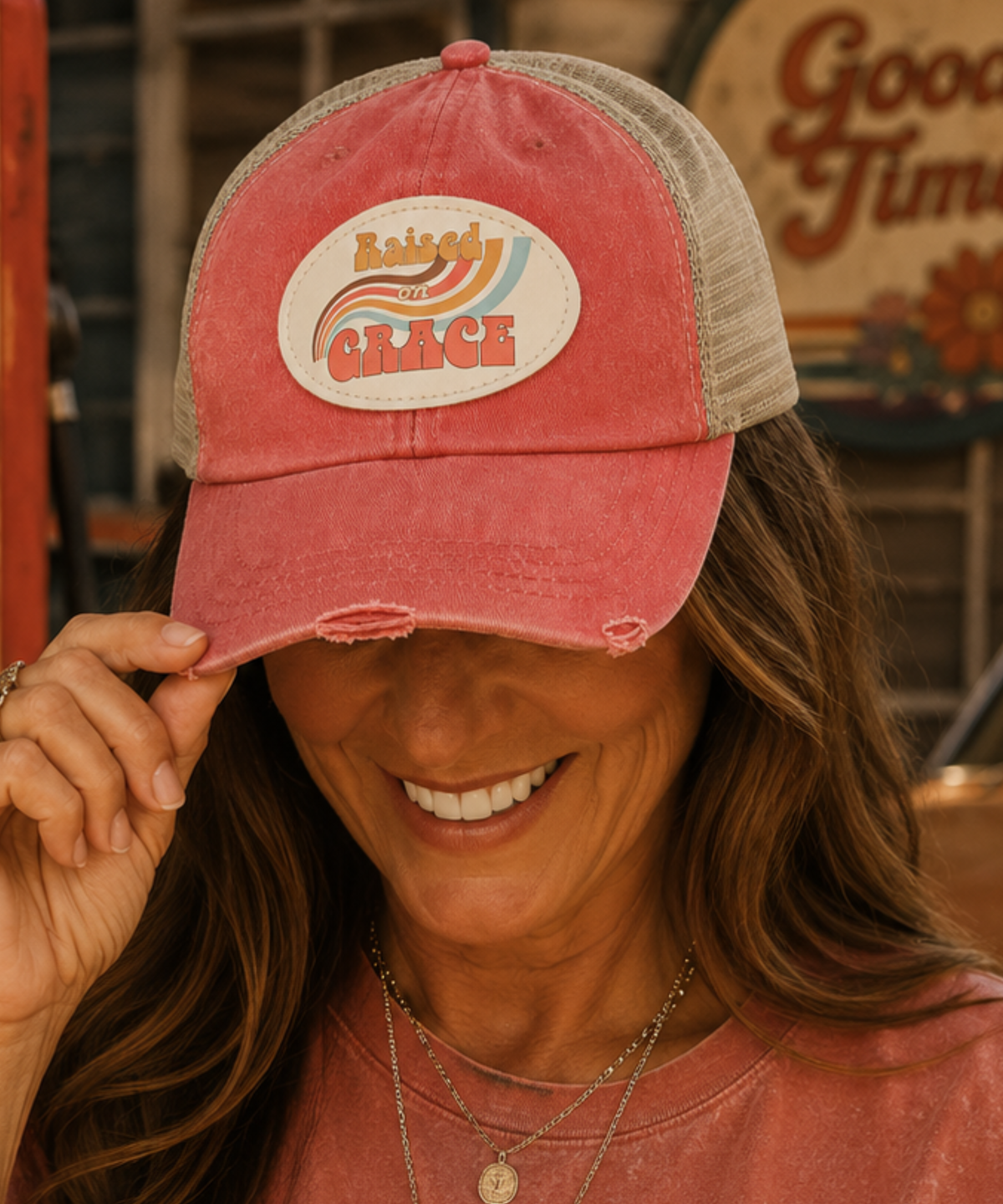 A woman wearing a distressed coral trucker hat with a beige mesh back and a circular 'Raised by Grace' patch on the front, smiling while holding the brim.