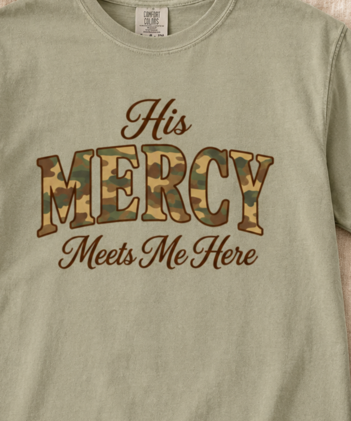 A close-up of a khaki t-shirt featuring the text 'His MERCY Meets Me Here' printed on the chest. The word 'MERCY' is filled with a classic camouflage pattern, while 'His' and 'Meets Me Here' are in a dark brown cursive font.