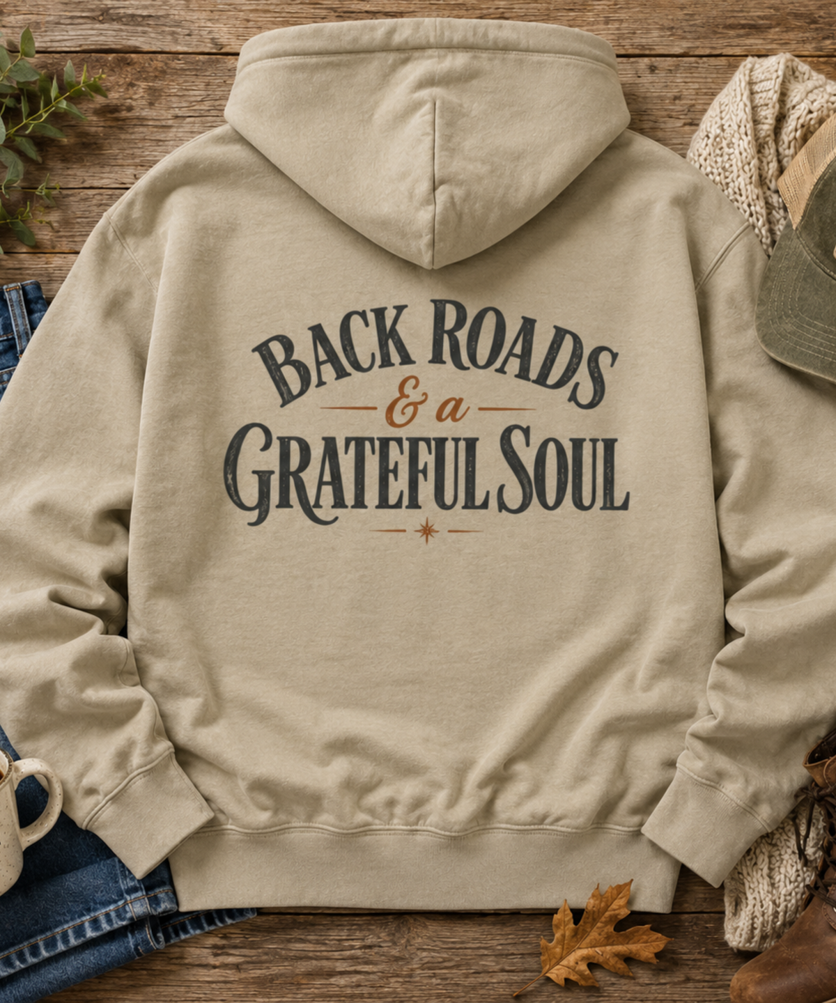 A gray pullover hoodie laid flat on a rustic wooden surface, shown from the back with the distressed text 'BACKROADS & a GRATEFULSOUL' printed in dark gray and brown lettering. The hoodie is styled with jeans, a knit sweater, a hat, a coffee mug, and autumn leaves.