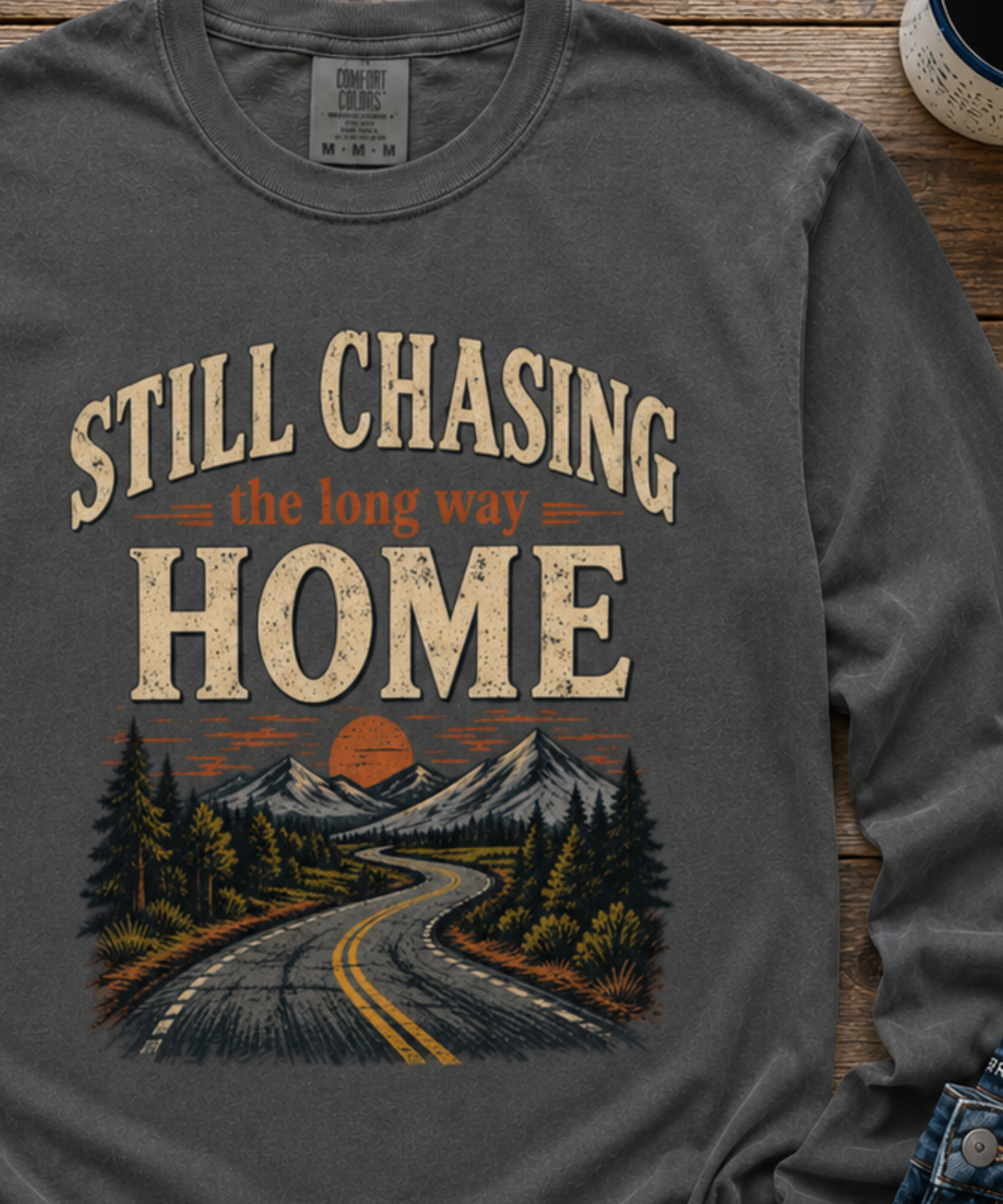 A close up of a pepper colored long-sleeve t-shirt with a vintage-style graphic print on the front. The graphic features the text 'STILL CHASING the long way HOME' in distressed beige and orange lettering above an illustration of a winding road leading through pine trees and mountains toward a sunset.