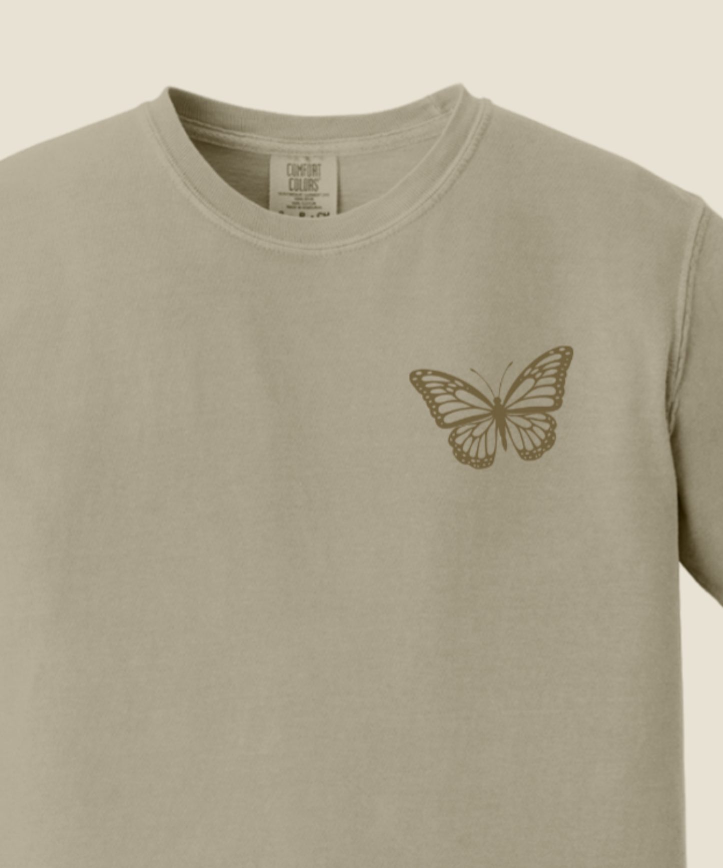 A close-up of a sandstone crewneck t-shirt featuring a small brown butterfly graphic printed on the left chest.