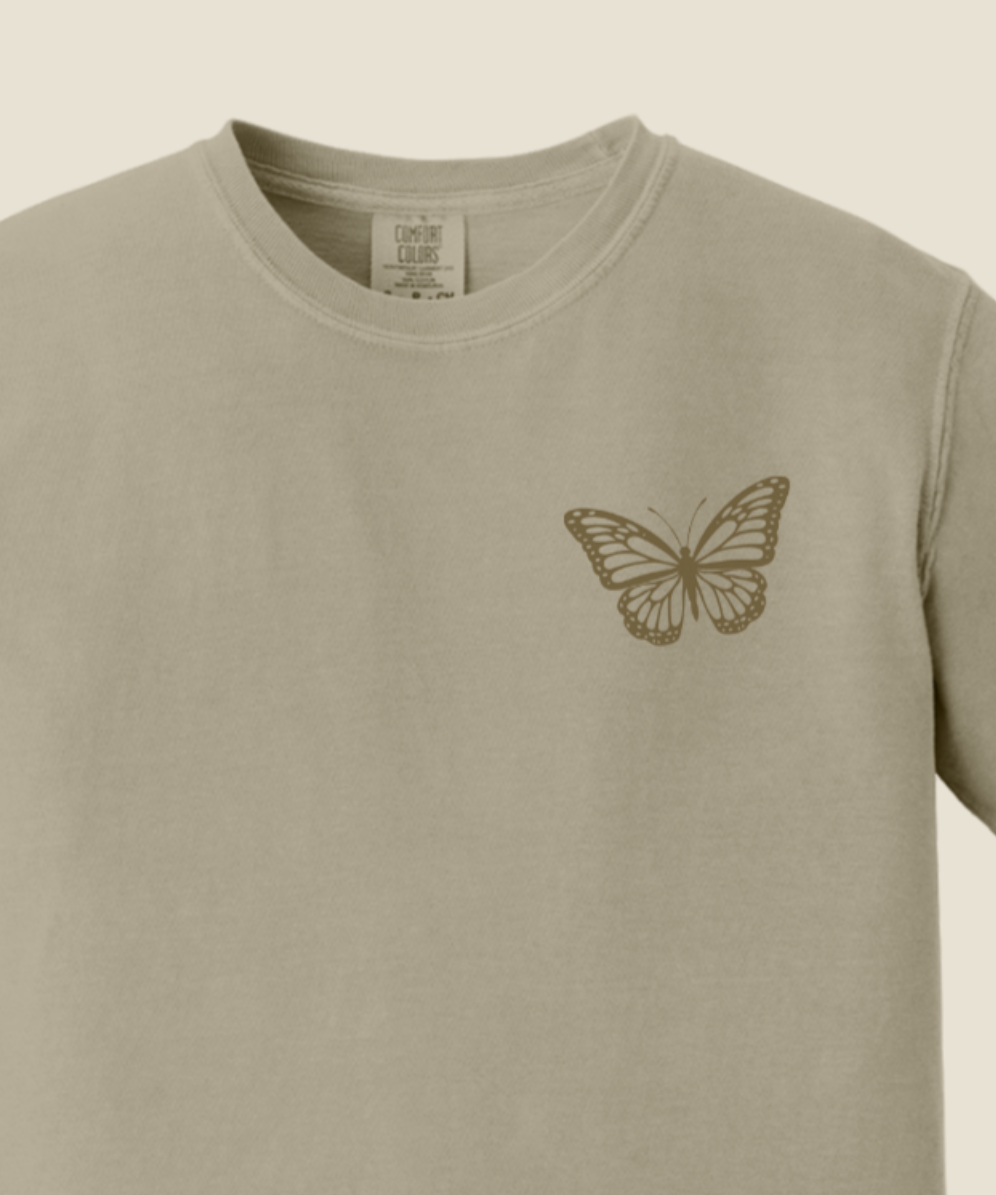 A close-up of a sandstone crewneck t-shirt featuring a small brown butterfly graphic printed on the left chest.